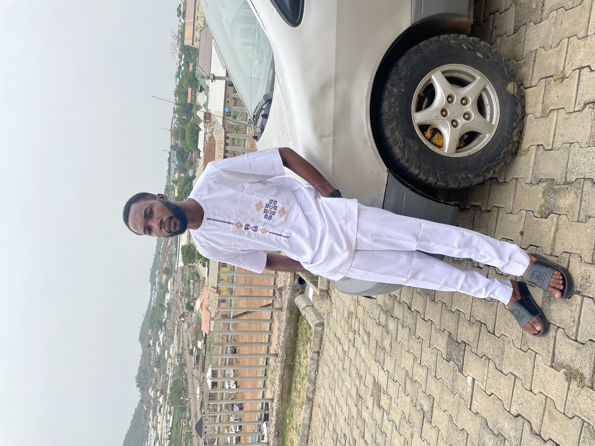 A man stands next to a silver car in Lokoja, Nigeria. He is wearing a white outfit with traditional embroidery on the chest pocket and sleeves and black Gucci slides. The car's front tire is visible behind him. The location appears to be an elevated area or rooftop with a view of the city of Lokoja, Nigeria. Buildings and surrounding hills are visible in the background under an overcast sky. The ground is paved with interlocking concrete pavers.