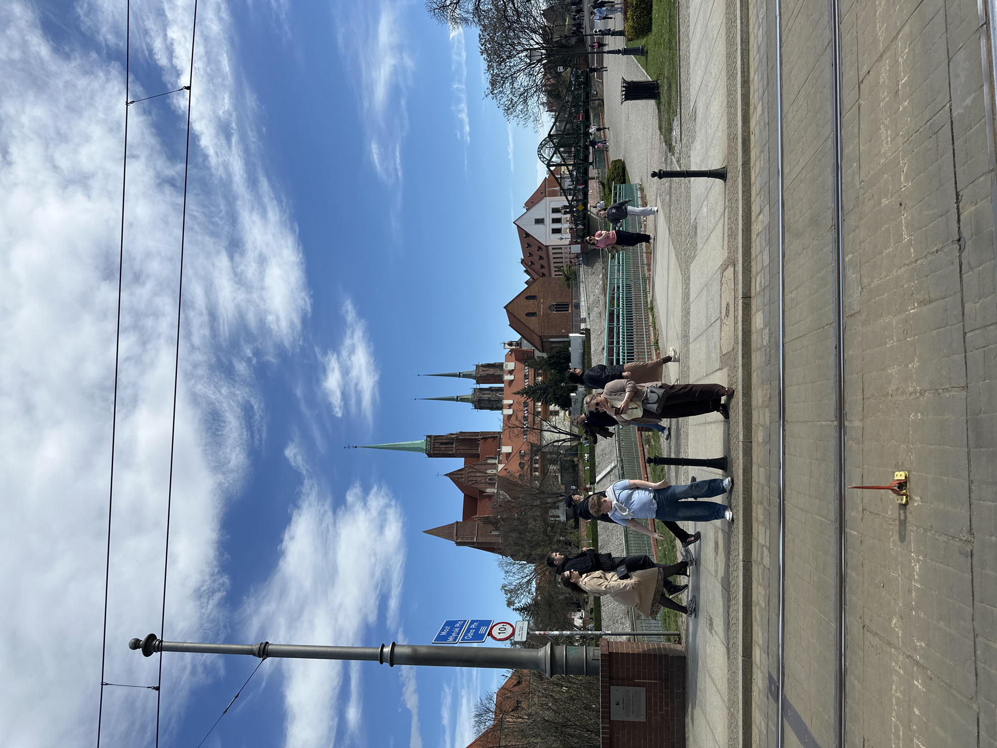 The image shows a view of Wrocław Cathedral in Wrocław, Poland, under a blue sky with scattered clouds. Several people are walking along a sidewalk parallel to tram tracks. The sidewalk is paved with gray stone. The foreground includes a street lamp and a signpost. The cathedral's spires are visible against the skyline. A bridge can be seen in the background.