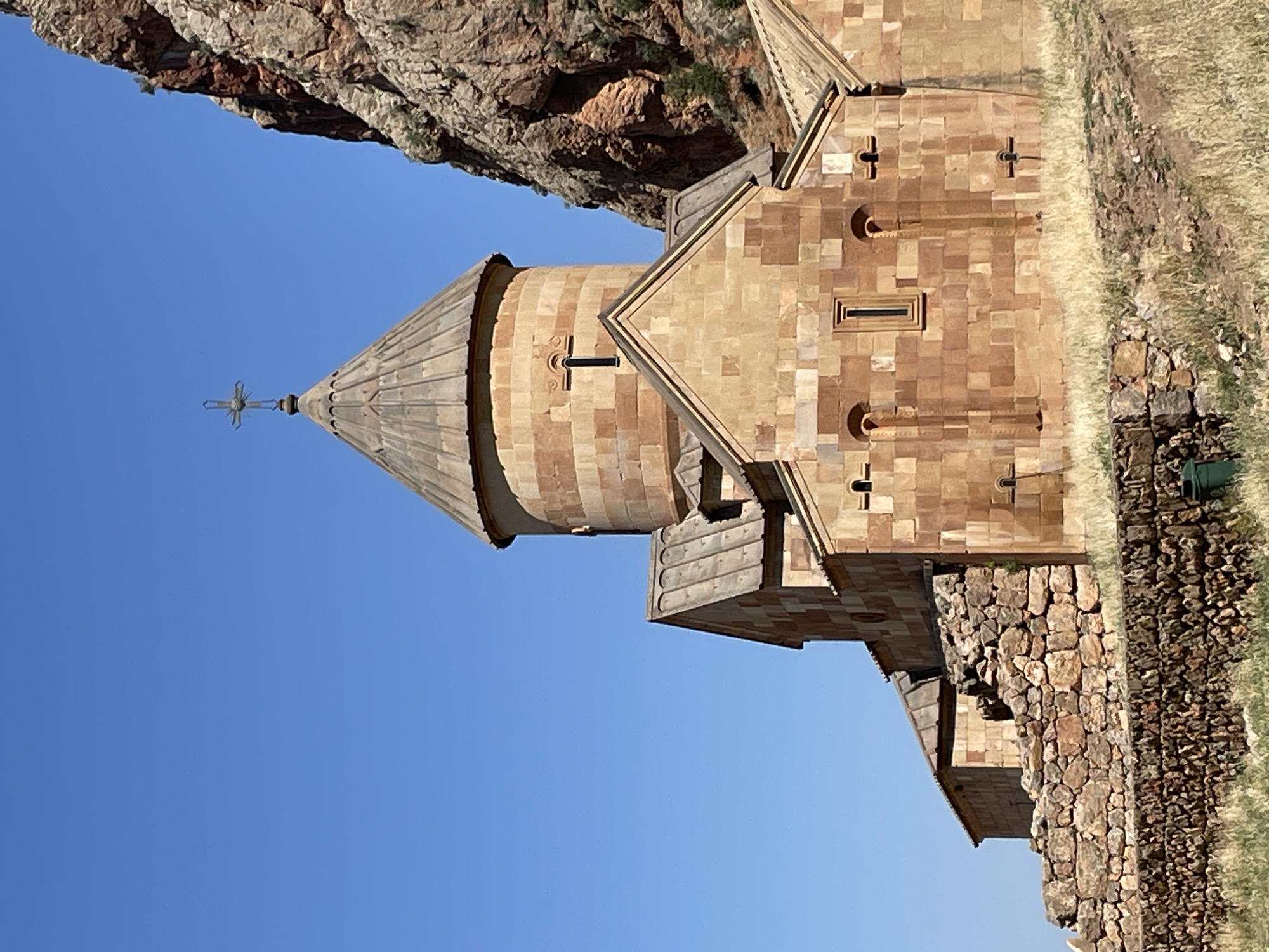 The image shows a stone church, likely Noravank Monastery, located in the city of Areni, Armenia. The church is constructed of reddish-orange stone blocks and features a conical roof topped with a cross. A large, rocky cliff face dominates the right side of the frame, casting a shadow over part of the church. The foreground is a dry, grassy landscape with low stone walls. The sky is clear and blue. There are no people or animals visible in the image.