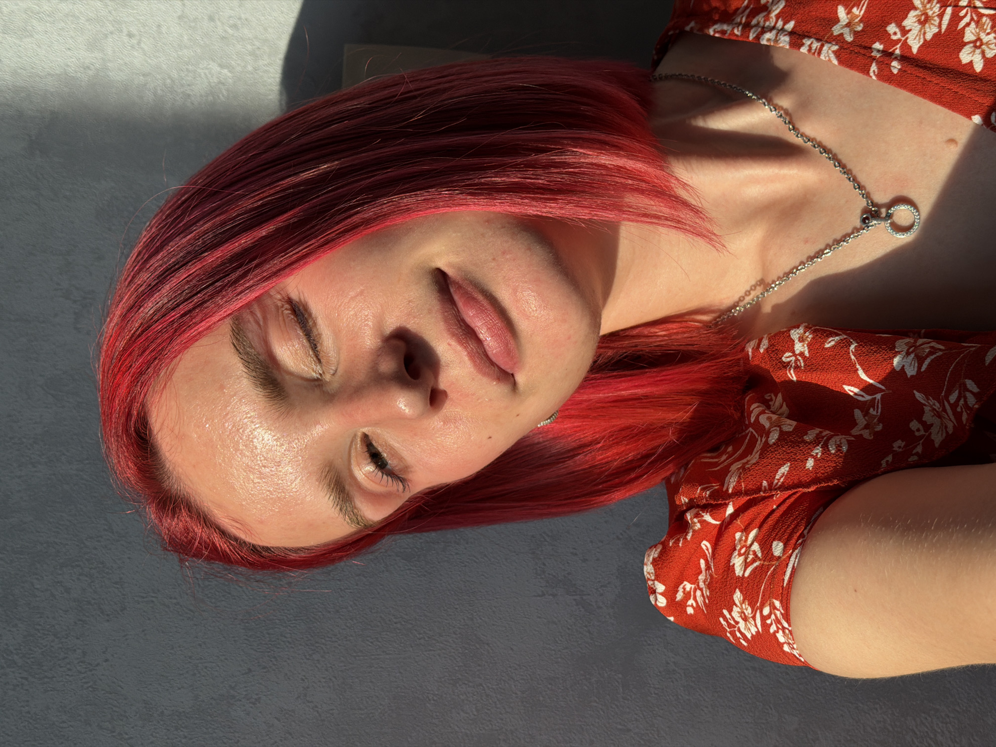 A young woman with bright pink hair and a red floral print dress is shown in a close-up portrait. She is looking towards the right with her left eye closed and a slight smile on her lips. The sun is casting shadows on her face and the wall behind her, which appears to be textured and gray. She is wearing a silver necklace with a circular pendant. The image is taken indoors, with the sunlight suggesting it is daytime. There is no information about the specific location within Chaikovsky, Russia.