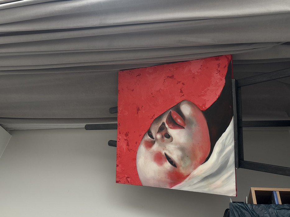 An oil or acrylic painting on canvas is positioned centrally on a dark-colored studio easel, set against a light-colored wall and light gray draped curtains. The painting depicts a stylized portrait of a human face with a pale complexion, featuring prominent red blush-like marks on the cheeks and forehead. The eyes are mostly closed or downcast, and the lips are full, painted a vibrant red. The head is tilted to the viewer's left, with a segment of white drapery visible around the lower-left portion of the face. The background of the painting is a textured, vibrant red. To the immediate left of the easel, a dark multi-tiered shelving unit is partially visible, holding a crumpled dark object, possibly fabric, and other indistinct items. The specific location within Krasnoyarsk, Russia, cannot be determined from the visual information provided in the image.