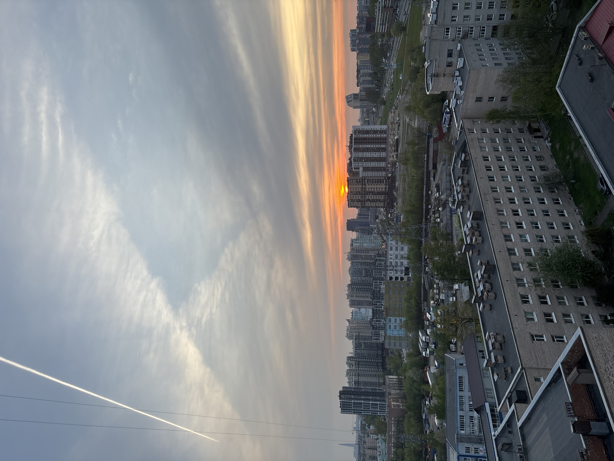 The image depicts a cityscape at sunset. The sky is streaked with orange and yellow light, interspersed with grey and white clouds. Buildings of varying heights line the horizon, partially obscuring the setting sun. The architecture is modern, with high-rise apartment blocks and commercial buildings dominating the skyline. Below, there are green trees and vehicles, suggesting a populated urban area. In the lower left corner, there are contrails from an airplane. A powerline is seen in the lower left corner. The photograph is taken from an elevated position, providing a wide, panoramic view of the city.