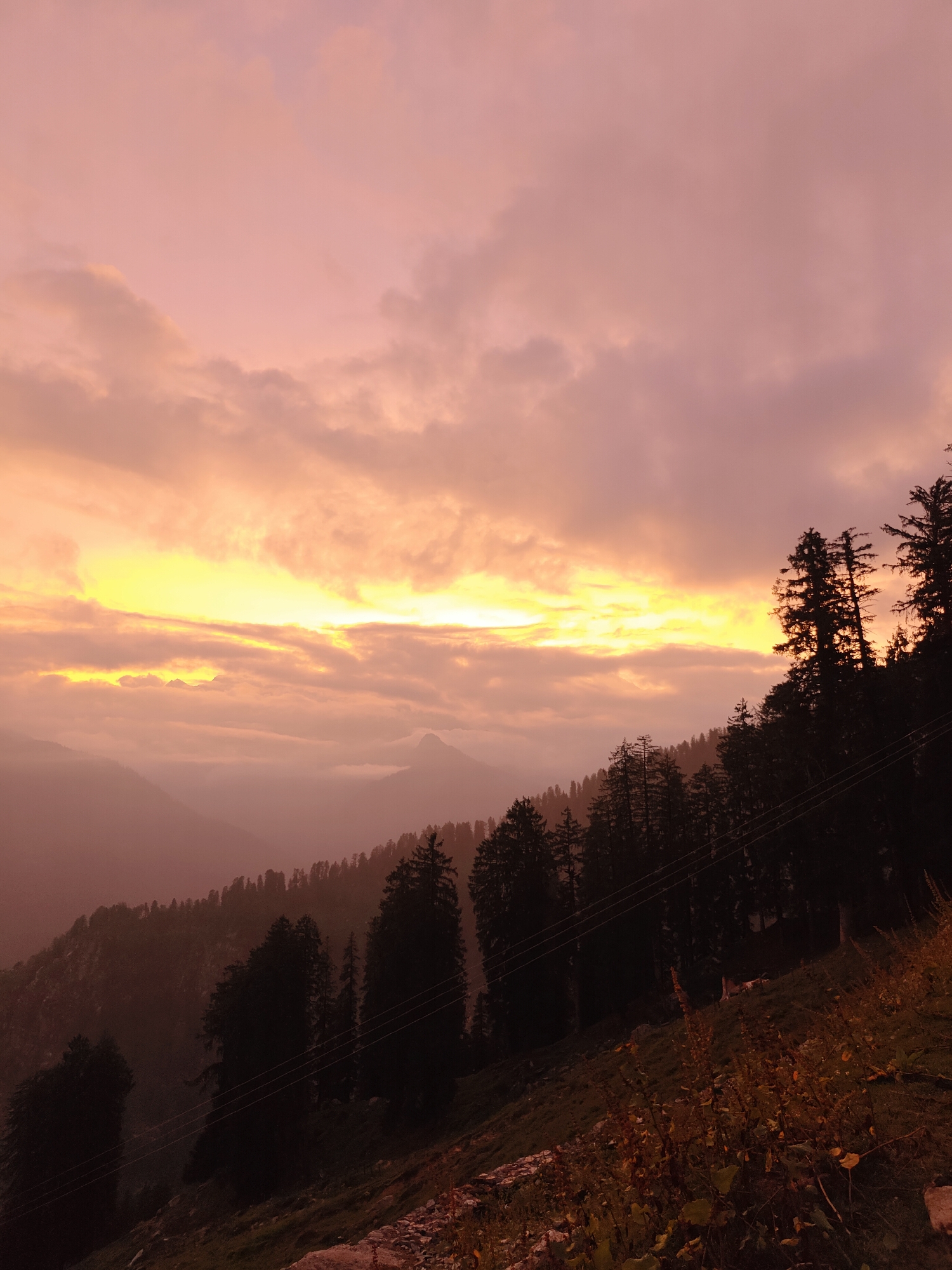 A sunset over a mountain landscape in Prini, India, is depicted. The sky is filled with clouds in shades of pink, orange, and yellow, with the sun's light breaking through the clouds in the distance. Silhouetted trees and mountains create a layered effect in the foreground and background. The foreground shows a grassy slope with scattered rocks and vegetation. The overall mood is serene and atmospheric.