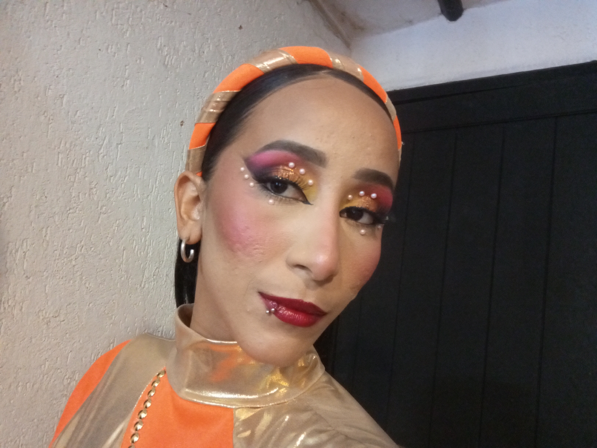 A person with orange and gold clothing and an orange headband with gold trim is shown in a close-up, indoor shot. The person has dramatic makeup including red lipstick, heavy pink and yellow eyeshadow with small white beads near the eyes, and prominent blush. They have a lip piercing on the lower lip and are wearing hoop earrings. The background consists of a white wall on the left and a dark wall, possibly a door, on the right. The location and city of Porlamar, Venezuela cannot be determined from the provided image.