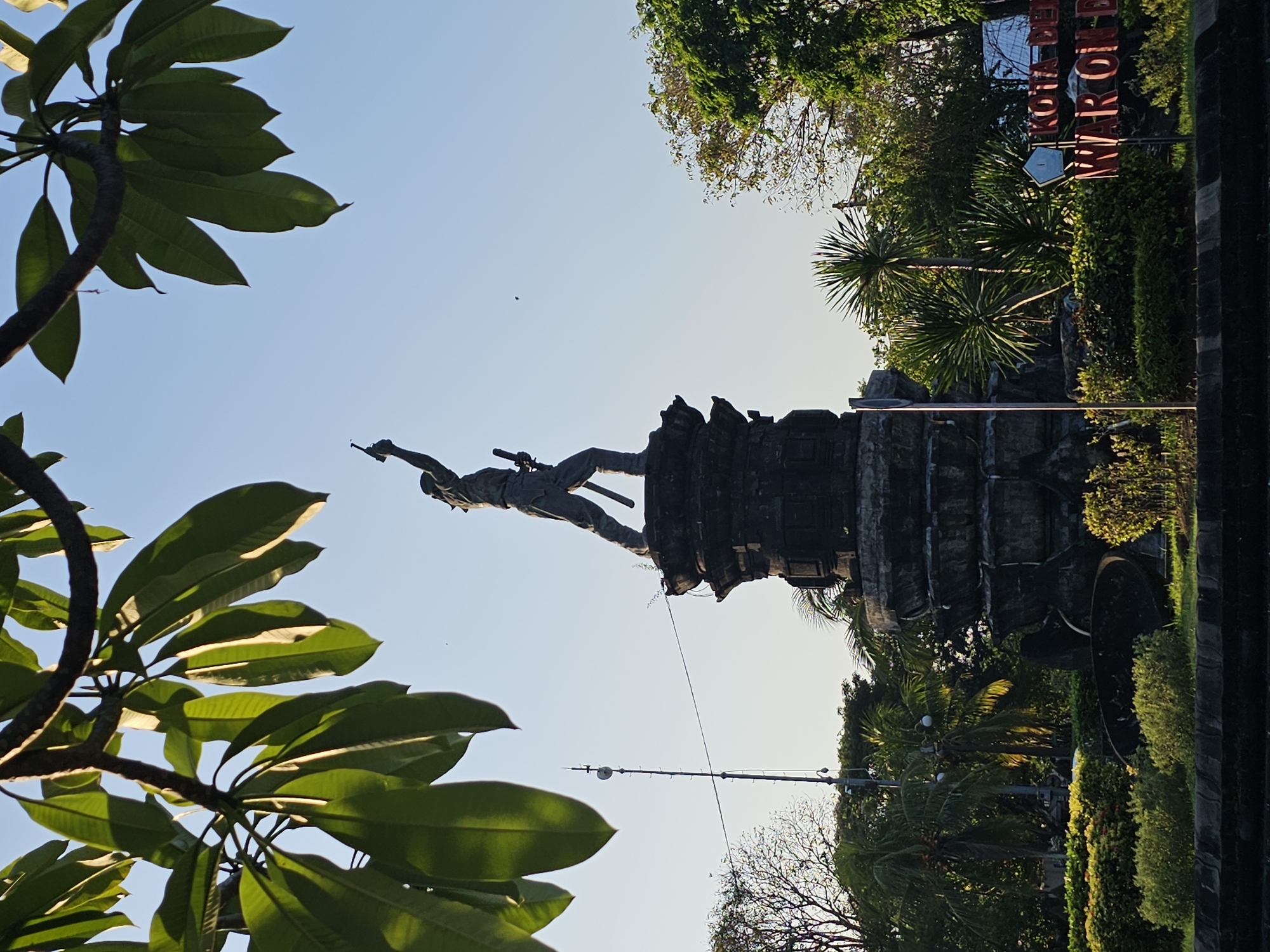 The image depicts the Puputan Badung Monument in Denpasar, Bali, Indonesia. The monument features a statue of a figure holding a rifle aloft, standing on a multi-tiered stone pedestal. The statue is silhouetted against a clear blue sky. Lush green trees and foliage surround the monument, with some frangipani leaves hanging into the foreground from the top left of the frame. A telecommunications tower is visible to the left of the monument, and in the lower right corner, a sign with red lettering reads "KOTA DENPASAR" and "PUPUTAN BADUNG". The scene appears to be captured during daylight hours with soft, warm lighting.