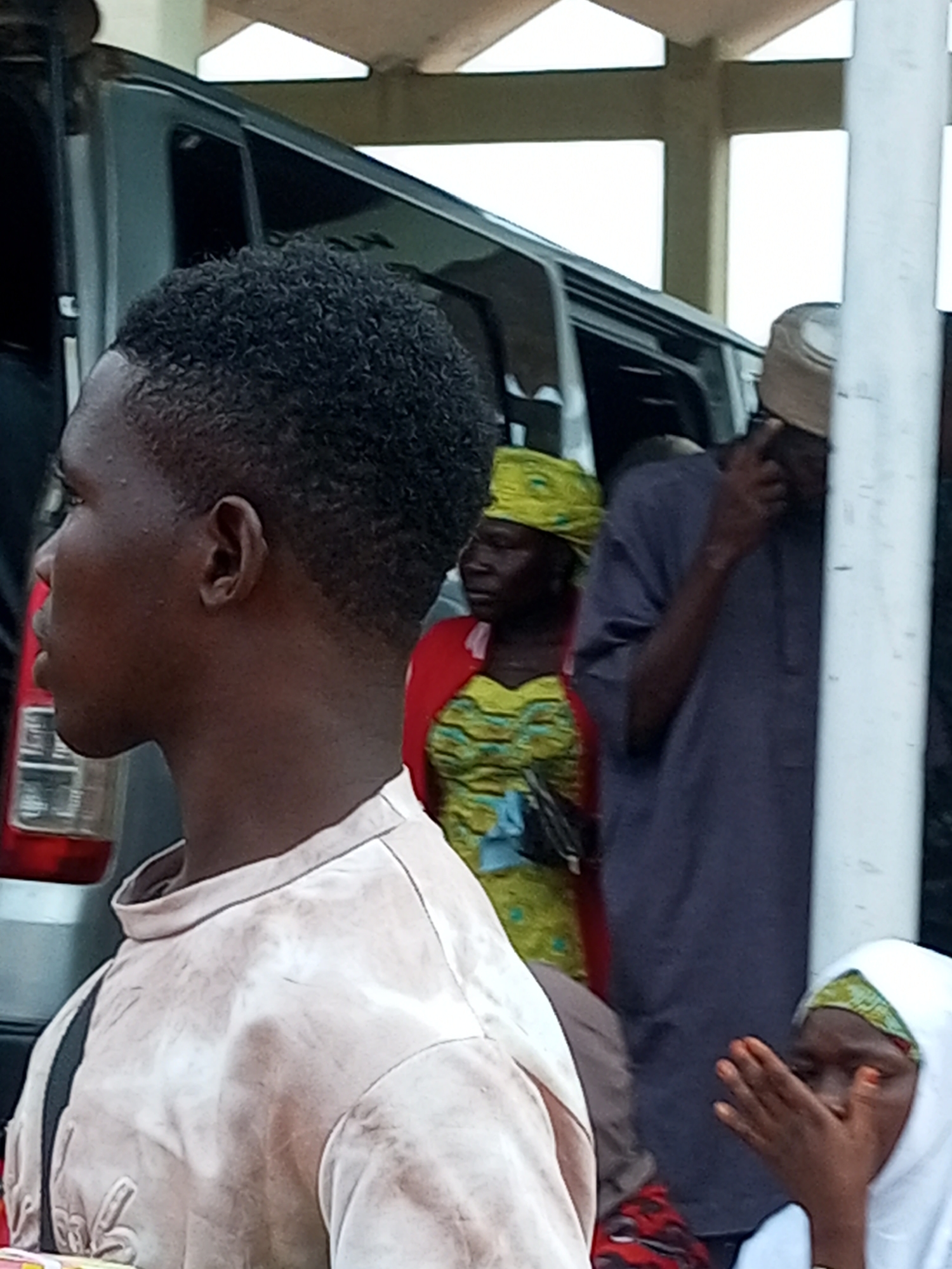 In Gombe, Nigeria, a group of individuals stand near an open vehicle. A young man with dark, curly hair is in the foreground, seen in profile, wearing a light-colored t-shirt. Behind him, a woman in a vibrant yellow and green patterned dress and red headscarf is visible. To the right, a person in a dark grey tunic and a cap appears to be speaking on a mobile phone. Another individual, partially obscured, wears a white hijab and has their hands covering their face. The background shows the side of a van with a red taillight and a white pillar.