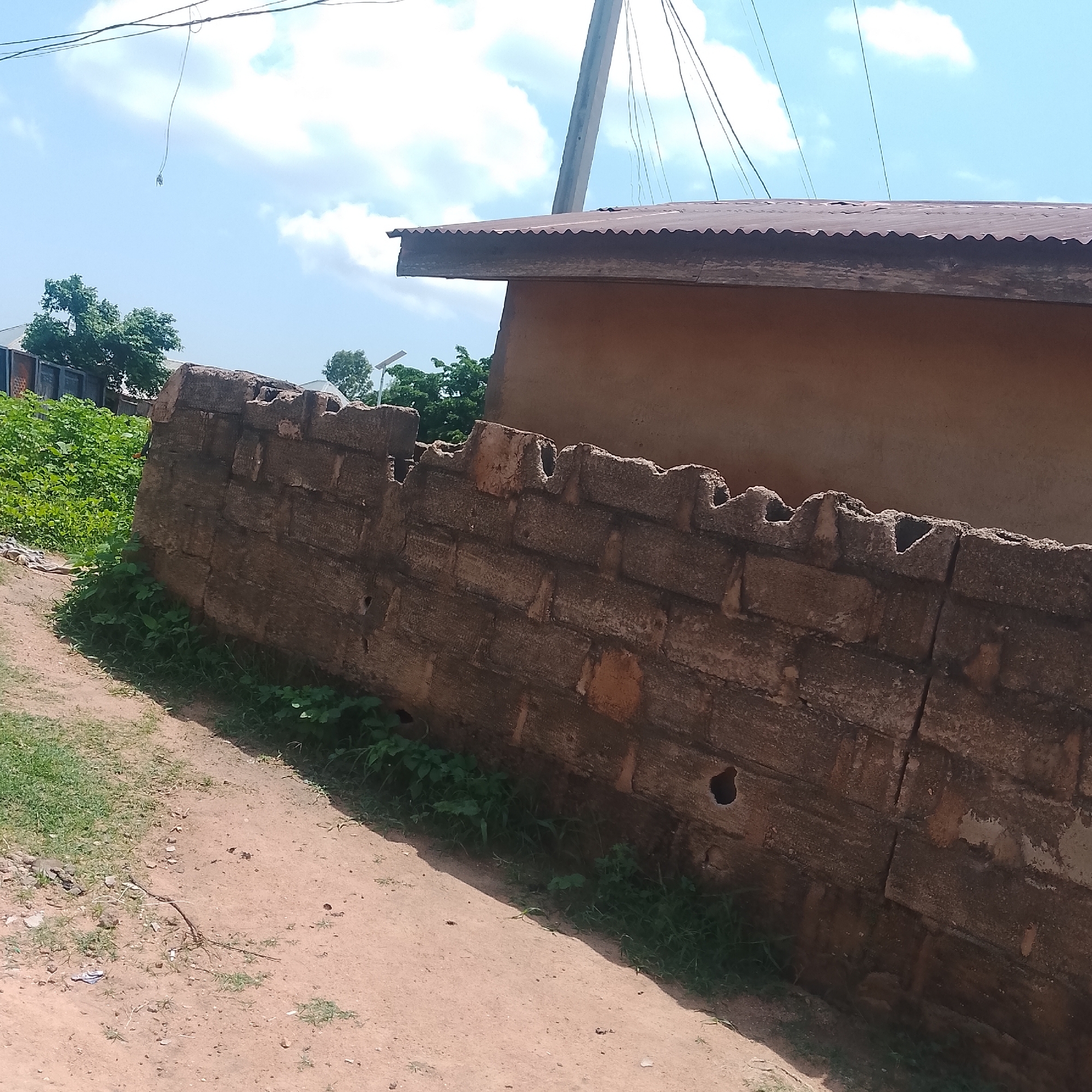 This media file captures an outdoor scene in Jalingo, Nigeria, under a bright, partly cloudy sky, suggesting daytime. The focal point is a weathered, rough brick wall that dominates the foreground and middle ground, extending diagonally across the frame. The wall is constructed of irregularly shaped and textured bricks, with visible gaps and holes, indicating age and perhaps disrepair. Lush green vegetation grows along the base of the wall and spills onto the sandy, dirt path that runs alongside it.
Behind the wall, a building with a corrugated metal roof is partially visible. The building's walls appear to be made of earth or mud, with a muted, tan color. Above the building, utility wires stretch across the sky, connected to a thin, tall pole. In the background, more greenery and hints of other structures can be seen, suggesting a semi-urban or rural environment. A solar panel is visible on a pole in the distance, adding a modern element to the otherwise rustic setting. The overall impression is one of a warm, sunny day in a community where traditional building materials and practices are still in use, interspersed with some modern infrastructure. There is no visible text in the image.