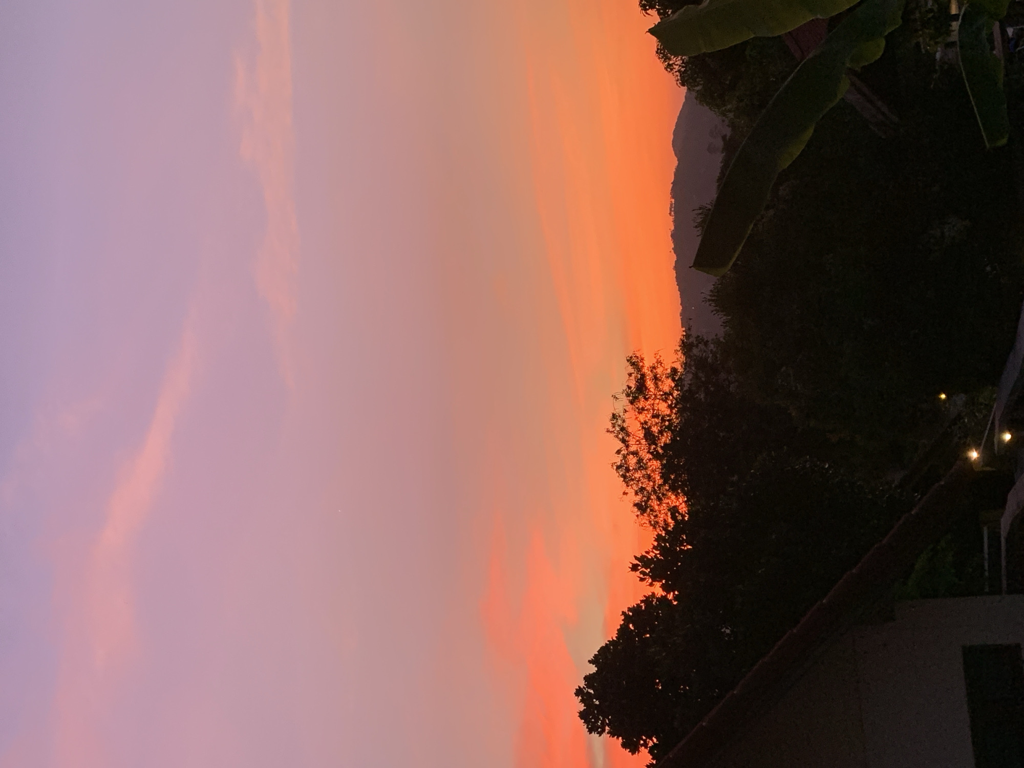 Captured in the city of บ้านบางรักษ์, Thailand, the image portrays a vibrant sunset. The sky transitions from a deep violet on the left to a vivid orange in the center, eventually meeting the silhouettes of trees and possibly a mountain in the right. The right side of the image is darker, where trees and a rooftop are visible. A few small lights can be seen, hinting at human habitation.