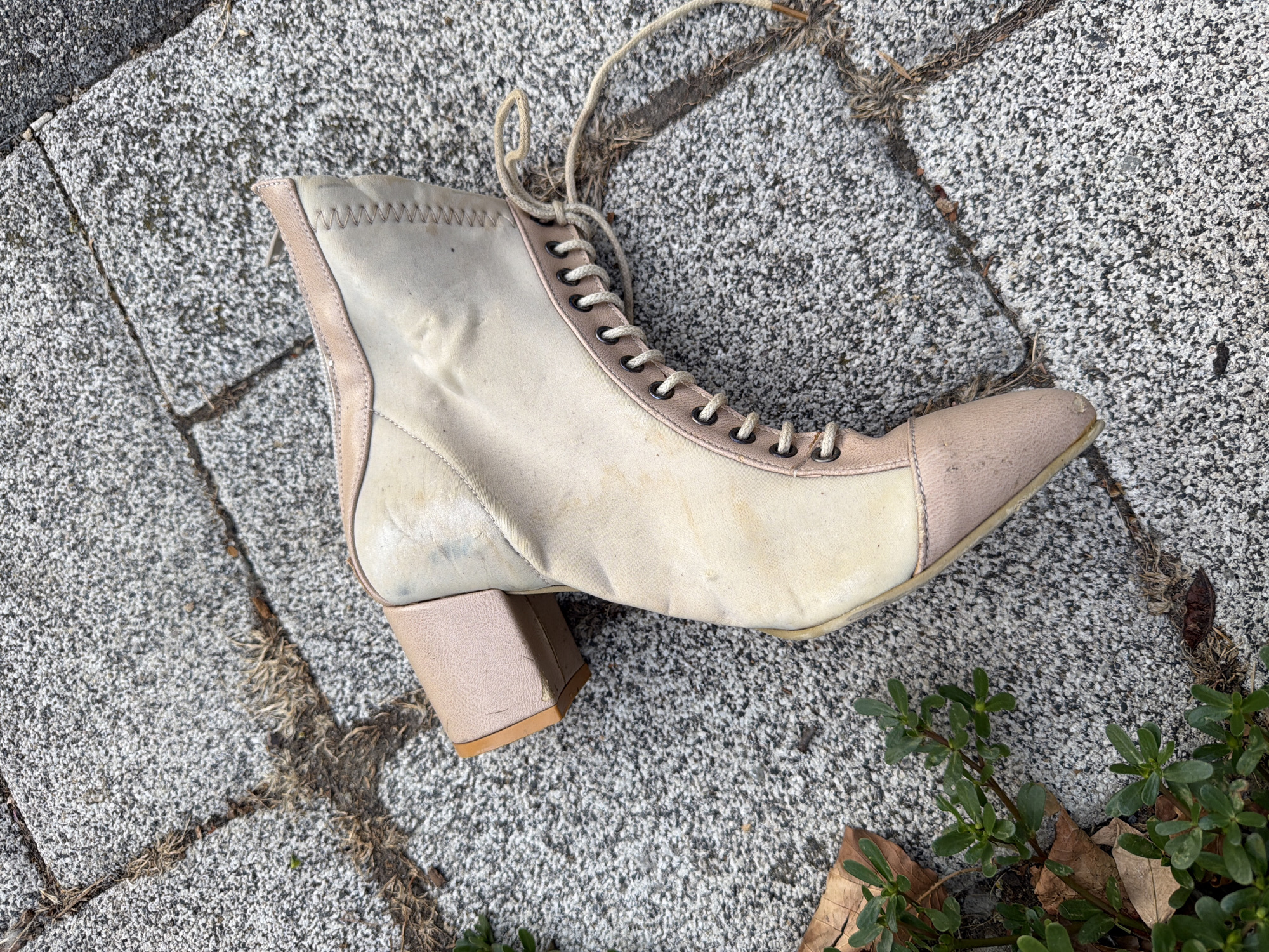A single, worn, cream and light pink ankle boot lies on a cracked, grey stone sidewalk. The boot has a chunky heel and laces that are partially undone. There is a small patch of green plants and dried leaves to the left of the boot. The scene is photographed from a high angle. The location is Üsküdar, Turkey.