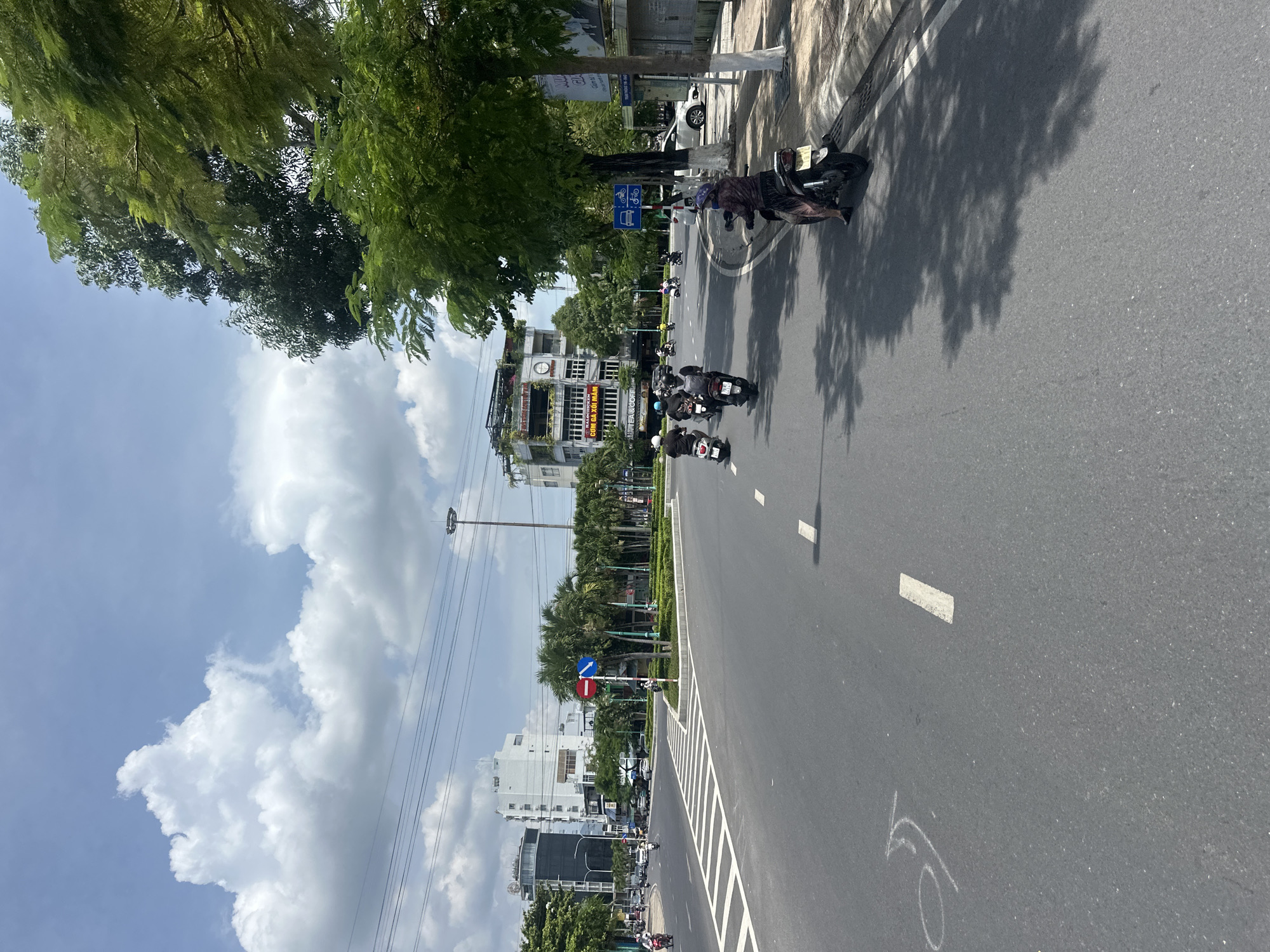 The image shows a wide, paved road in Bac Nha Trang Ward, Vietnam, under a partly cloudy sky. Several motorbikes are visible on the road. In the foreground, a person on a motorbike is stopped on the right side of the road. Further ahead, a group of motorbikes are traveling in the same direction. The road has white lane markings. On the left side of the road, there are traffic signs indicating a turn and a no-entry zone. In the background, there are several multi-story buildings, trees, and streetlights. The overall scene depicts a typical urban streetscape in Vietnam with prevalent motorbike traffic.