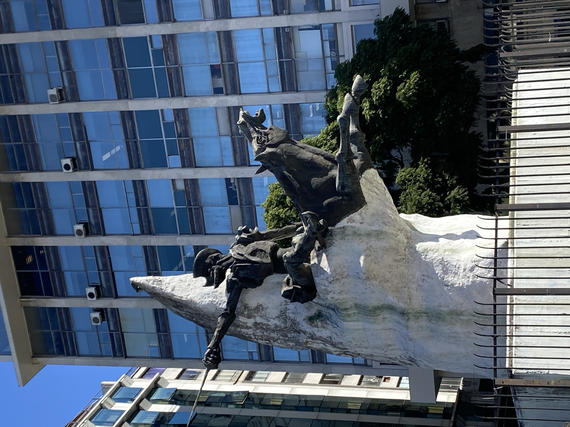 The image displays a prominent bronze equestrian sculpture set against a backdrop of modern buildings on a bright, sunny day. The sculpture, positioned centrally, features a figure on a rearing horse atop a large, textured white base that evokes a wave or rocky outcrop. Both the horse and rider exhibit a dynamic, heroic pose, rendered in dark, weathered bronze.
In the background, a tall, multi-story building dominates, characterized by its grid-like facade of blue-tinted, reflective glass windows. Several white external air conditioning units are mounted on its exterior. To the right of the sculpture, and partially obscured, is another building with a series of horizontal architectural louvers or blinds. Lush green tree foliage is visible between the sculpture and the background structures, adding organic contrast to the urban setting.
The clear sky and distinct shadows indicate bright daylight conditions. No visible text is present in the image. The scene juxtaposes classical sculptural art with contemporary architectural design within what appears to be an urban environment, given the context of Buenos Aires, Argentina.