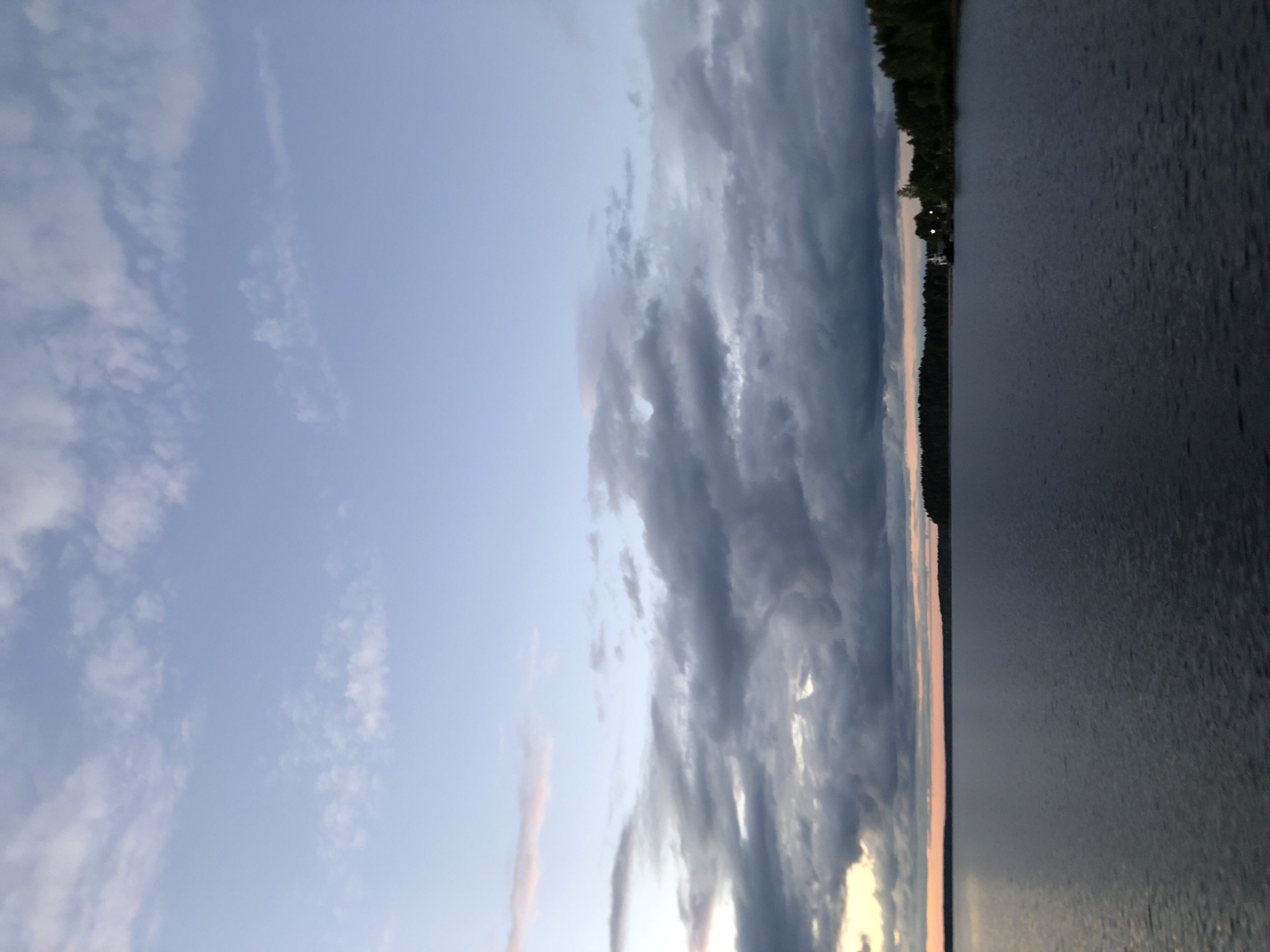 The image depicts a serene evening scene at Lake Ladoga in Svirstroy, Russia. The sky is a soft gradient of blue and pale pink, with scattered clouds in varying shades of grey and white. A calm body of dark water stretches across the foreground, reflecting the twilight sky. On the distant horizon, a dense line of dark green trees marks the edge of a wooded area. A small, white structure with a visible light can be seen nestled amongst the trees on the right side of the image, suggesting a settlement or a solitary building. The overall atmosphere is tranquil and peaceful.