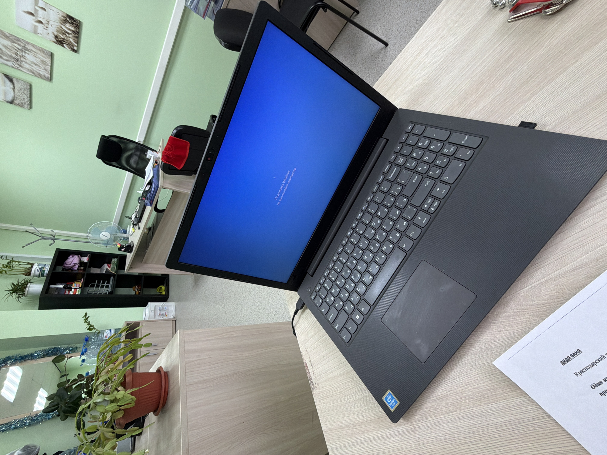 A slightly high-angle shot focuses on a black laptop placed on a light-colored wooden desk, likely in an office or study environment in Chaikovsky, Russia. The laptop screen is illuminated, displaying a Windows loading or preparation screen. The keyboard is clearly visible, and a printed document is positioned to the right of the laptop, with Russian text on it. A potted plant with green foliage is situated to the left of the desk. In the background, there is a shelf with various items, and a glimpse of a green wall with pictures hanging on it can be seen. A few chairs are also visible in the background. The overall lighting suggests an indoor setting with natural light.