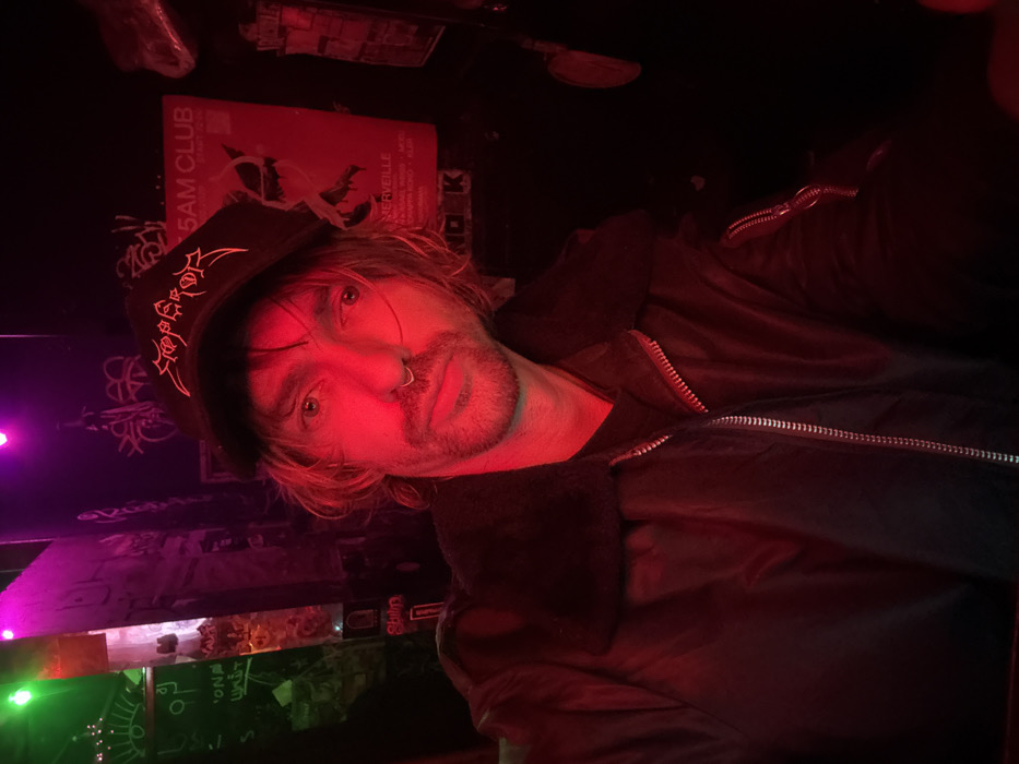 A man wearing a black jacket and hat poses in dim lighting. His hat features embroidered detailing. A ring pierces his nose. Red and purple light casts shadows across his face. Behind him is a wall with graffiti and posters, one of which reads "5 AM CLUB." The lighting creates a moody atmosphere.
