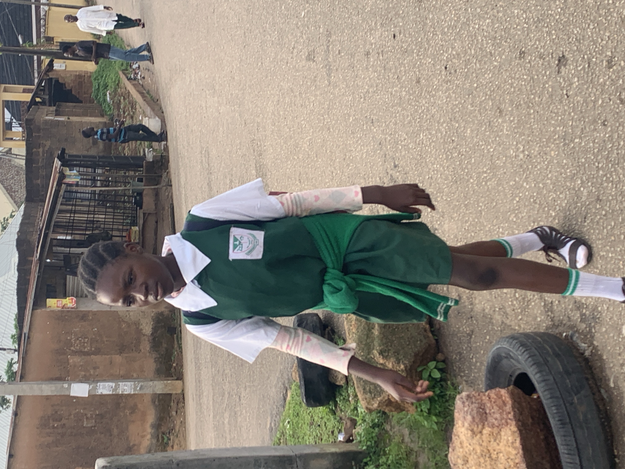 The image shows a young Black girl wearing a school uniform, consisting of a white collared shirt, a green vest, a green skirt, and white socks with green stripes. The uniform also includes a green sash tied around her waist. She has braided hair and is looking down. Her left arm is visible, adorned with a patterned sleeve in shades of pink and white. She is wearing black and white sneakers.
The girl is standing on a paved surface, likely a road, in an outdoor setting. To her right, there's a large, worn tire and a rock with some greenery growing near it. In the background, there are buildings with brick walls and metal fencing. Several other people are visible in the background, walking on the road. One person in the distance wears a white shirt and dark pants, while another person closer to the foreground wears a striped blue and black shirt and jeans. The overall scene suggests a street in a residential or commercial area.
The lighting suggests it could be daytime, possibly late morning or early afternoon, with diffused sunlight. The environment appears to be in a warm climate, as indicated by the clothing of the people and the presence of greenery. There are no clear indicators of specific weather conditions, but the scene appears dry.
One of the crops reveals a small emblem on the girl's school vest, featuring some text and symbols, but it is not legible enough to read. Another crop shows some posters or notices attached to a utility pole in the background.
The overall impression is a candid street scene, capturing the girl in her school attire within a typical Nigerian urban environment.