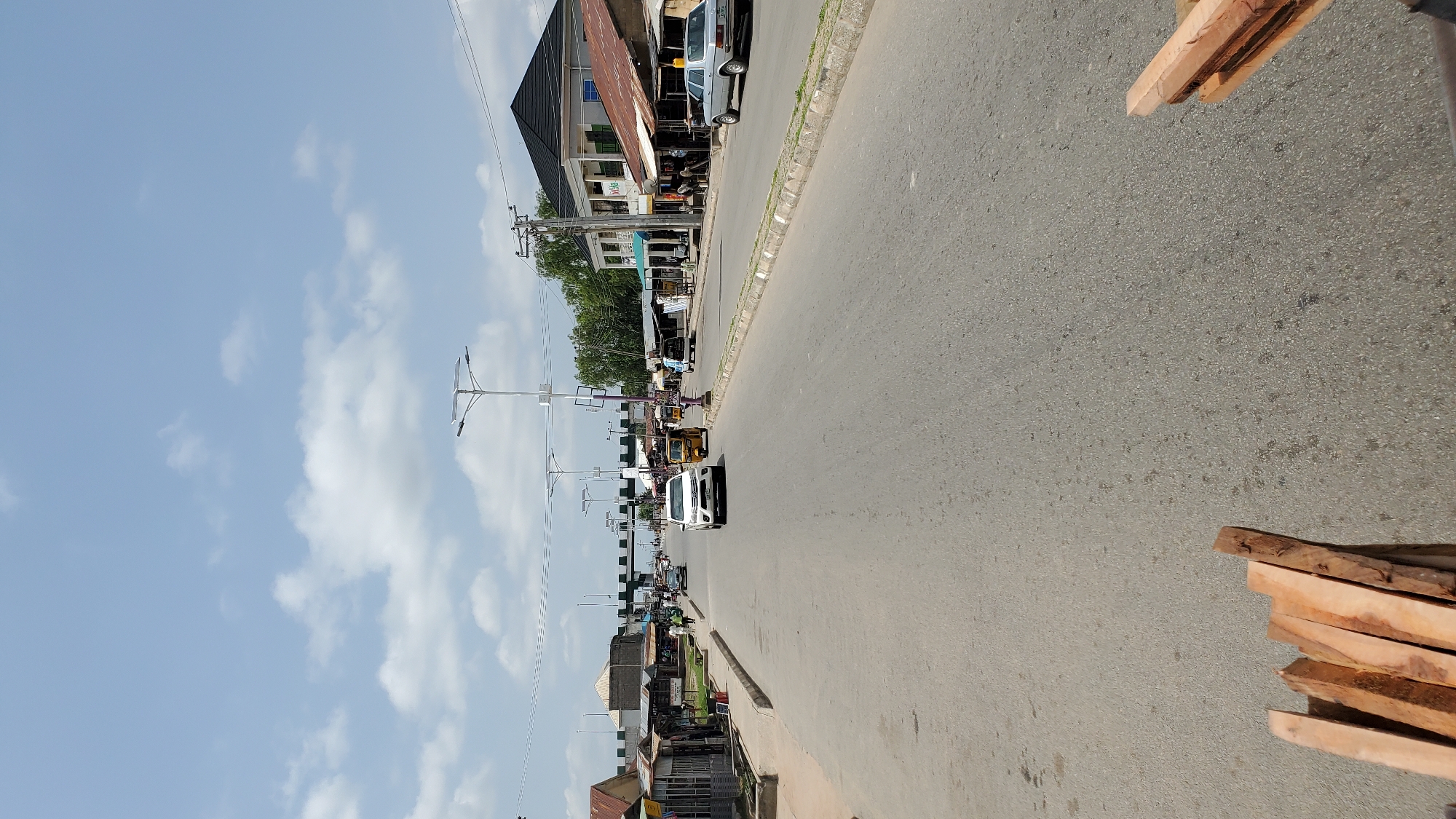 The image depicts a wide, paved road in Jalingo, Nigeria, under a bright, partly cloudy sky. The road is lined with various structures, including commercial buildings, shops, and residential homes. Several vehicles are present, including cars, a tricycle taxi, and a commercial van. The presence of solar-powered streetlights suggests modernization. In the foreground, a pile of lumber is visible, indicating potential construction or trade activities. The overall scene portrays a typical day in an urban environment, with people going about their daily routines.