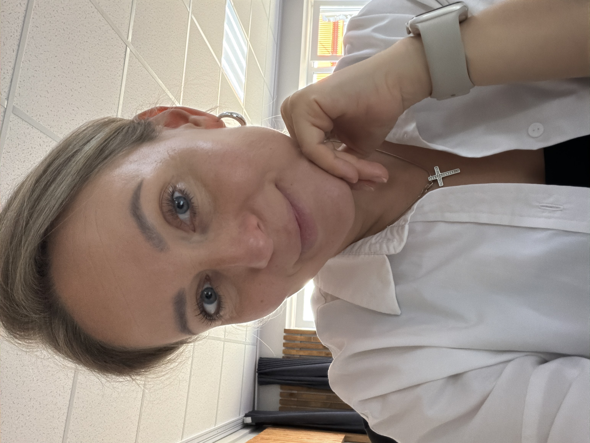 A person with short, light brown hair and blue eyes is looking directly at the camera. They are wearing a white collared shirt and a white smartwatch. A silver cross necklace is visible around their neck. Their hand is touching their face. The background includes a white tiled ceiling, a window with horizontal blinds, and a glimpse of what appears to be a wooden bench. Based on the request, this image was taken in Chaikovsky, Russia.