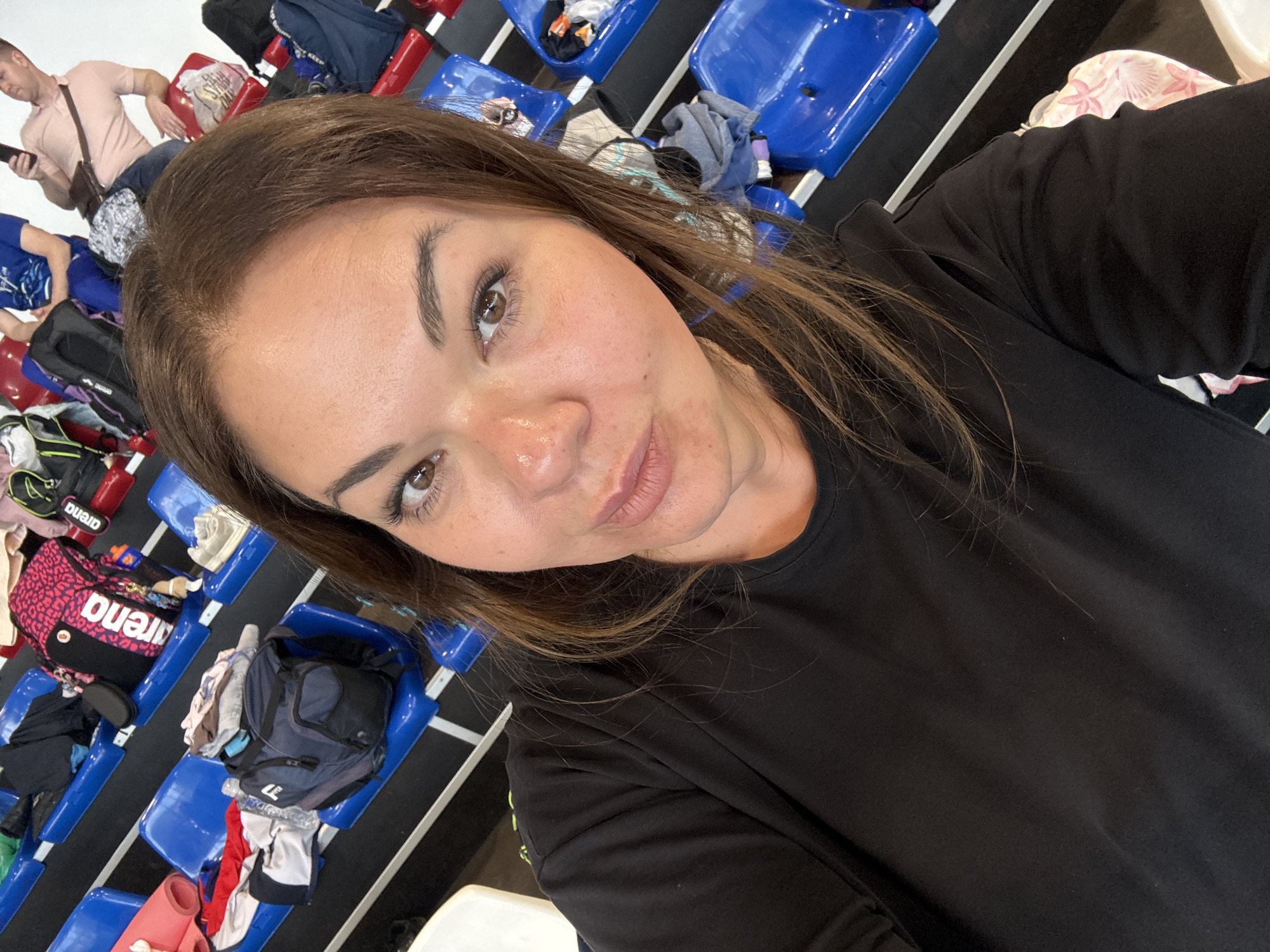 A medium-shot, slightly angled, selfie shows a woman with light brown hair and brown eyes wearing a black shirt. She is looking at the camera with a slight smile. Behind her are rows of blue and red stadium-style seats, some with backpacks and assorted items on them. A man in a pink shirt is visible in the background looking at his phone. Based on the presence of "Arena"-branded bags, the context of the image, and the stadium-style seating, the location may be related to a sporting event or competition. It is not possible to confirm the exact location or to state it is within Trebinje, Bosnia and Herzegovina, without further evidence.