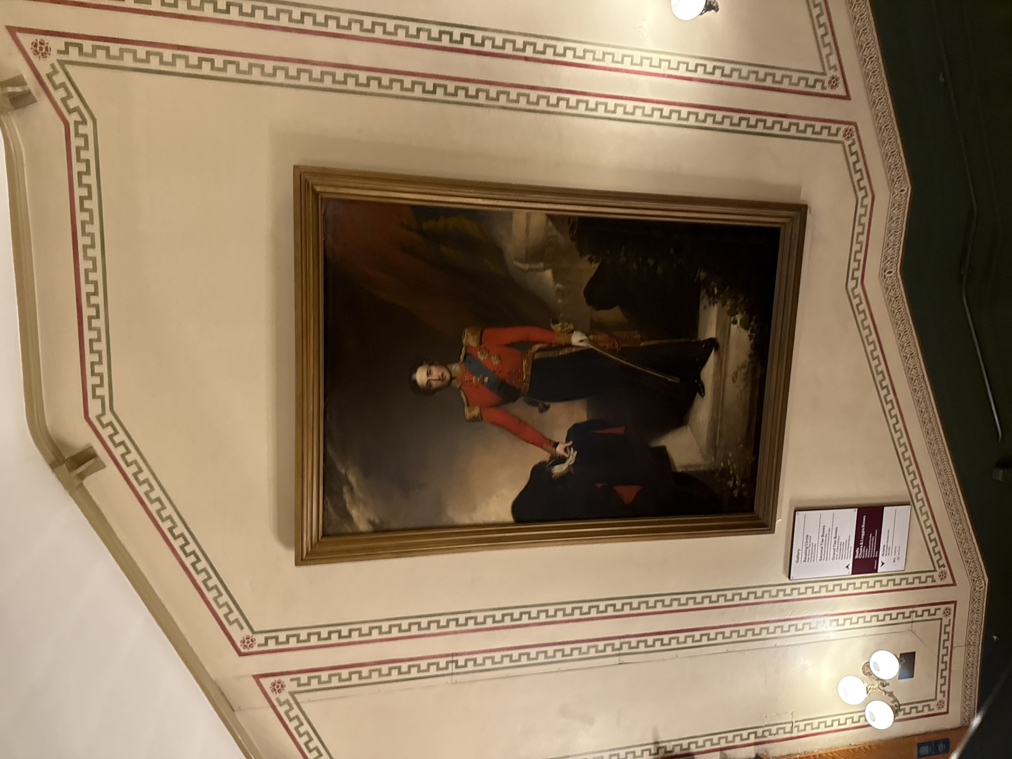 The image displays an interior view of a wall within the Royal Albert Hall, London, United Kingdom.
A large, vertically oriented oil portrait, framed in ornate gold, is centrally positioned on a light-colored wall. The painting depicts a full-length male figure standing, facing slightly to the viewer's right. He wears a red military-style uniform with gold epaulets, a blue sash, dark trousers, and black shoes. His left hand, clad in a white glove, rests on a dark cloak draped over an unseen support. His right hand holds a slender object, possibly a sword or cane, pointed downwards. The background of the portrait features a cloudy sky on the left and darker, indistinct landscape or architectural elements on the right.
The walls are a cream or off-white color, decorated with geometric borders. These borders feature a repeating double-line pattern, resembling a Greek key or meander, with alternating red and green lines. Red floral rosette motifs are present at junctions within the border design. A darker, possibly green, dado rail or lower wall section is visible at the bottom of the frame.
Below and to the left of the portrait, a brown rectangular sign with white text is mounted on the wall. The sign lists various seating sections and directions, including "Gallery," "Rausing Circle," "Second Tier Boxes," "Grand Tier Boxes," "Stalls," "Choir & Loggia Boxes," and "Arena," accompanied by directional arrows. A wall-mounted light fixture with three spherical bulbs, emitting a warm glow, is visible to the left of the sign. The ceiling, visible at the top of the frame, is also light-colored.