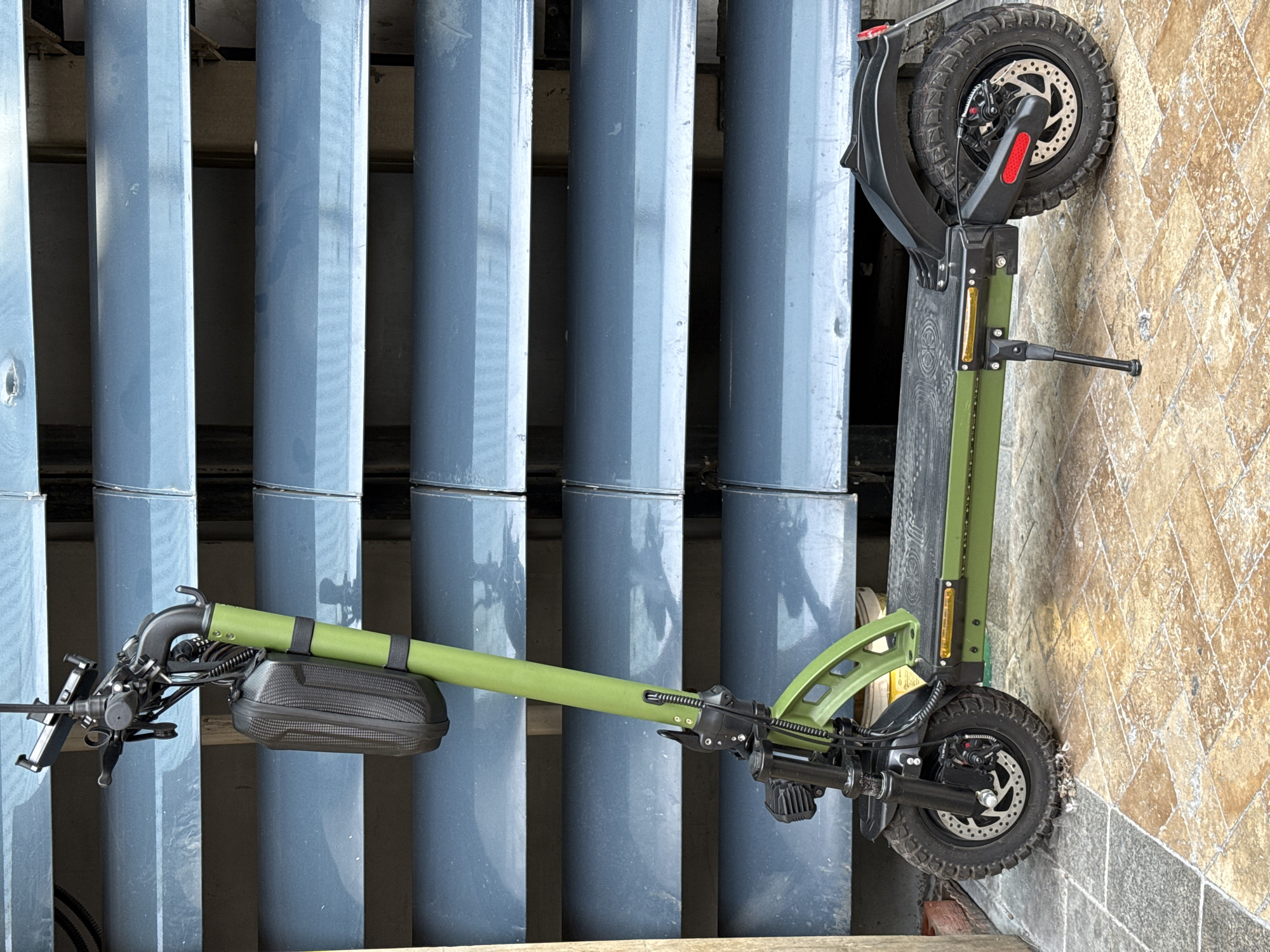 A green electric scooter is parked on a tiled walkway in front of a blue louvred structure. The scooter has a black bag attached to the handlebars and a kickstand extended. The scooter's front wheel has knobby tires and a disc brake. The rear wheel also has knobby tires and a disc brake, with a red reflector on the fender. The tiled walkway is made of light brown and grey stones arranged in a mosaic pattern. The background consists of horizontal blue slats, possibly part of a building or fence. The lighting suggests it is daytime. The image is taken from a medium-distance, eye-level perspective.