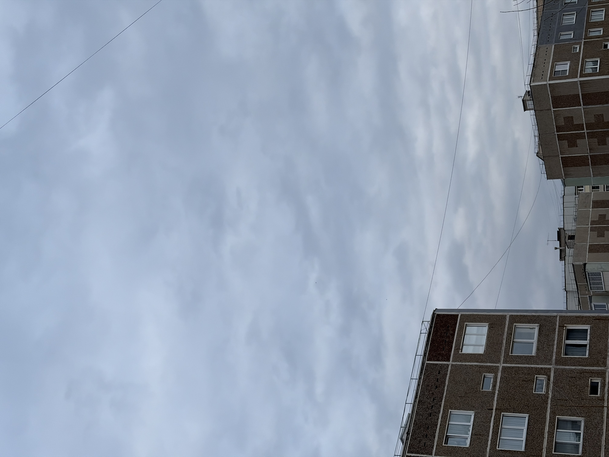 An upward-angled shot showcases an overcast sky, partially obscured by the tops of two Soviet-era apartment buildings. The buildings are constructed with prefabricated concrete panels, featuring a repetitive grid of windows. Exposed wiring stretches across the sky between the buildings. The location is likely Chaikovsky, Russia, based on the architectural style of the buildings which are typical of post-Soviet era urban planning.