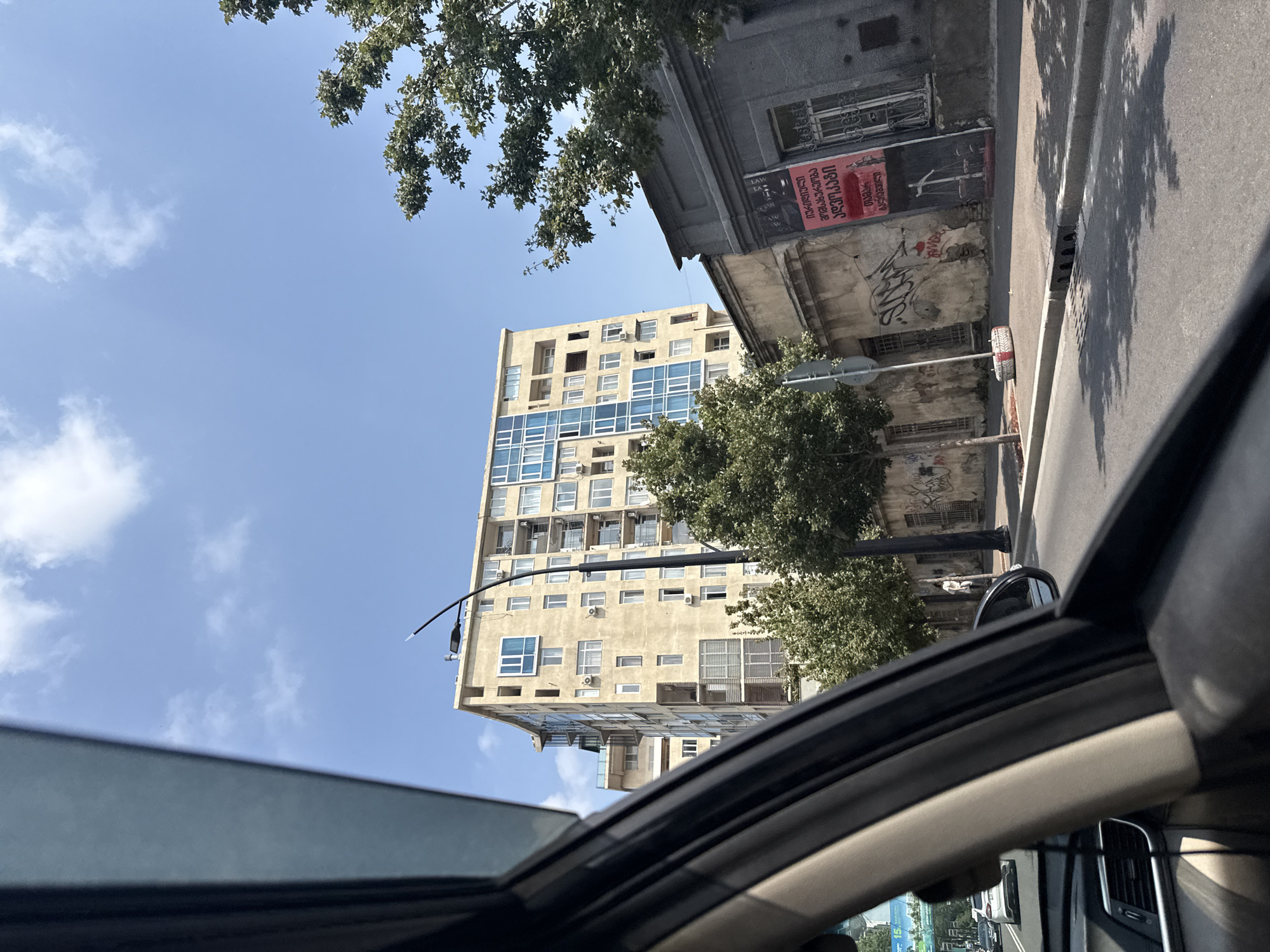 A low-angle exterior shot, taken from inside a vehicle, captures a view of a street scene in Tbilisi, Georgia. The sky is a bright blue with sparse white clouds. In the mid-ground, a tall, light-yellow apartment building with numerous windows and balconies dominates the frame. To the right of the apartment building is a lower, grey building with a weathered facade, featuring a large red poster with Georgian script and an illustration of scales. Graffiti is visible on the walls of both buildings. Trees with green foliage are interspersed throughout the scene. A street lamp stands prominently in front of the apartment building. A few cars are visible on the road in the background. The foreground shows the interior of a car, including the dashboard and the driver's side window frame. The street itself has a sidewalk and a curb.