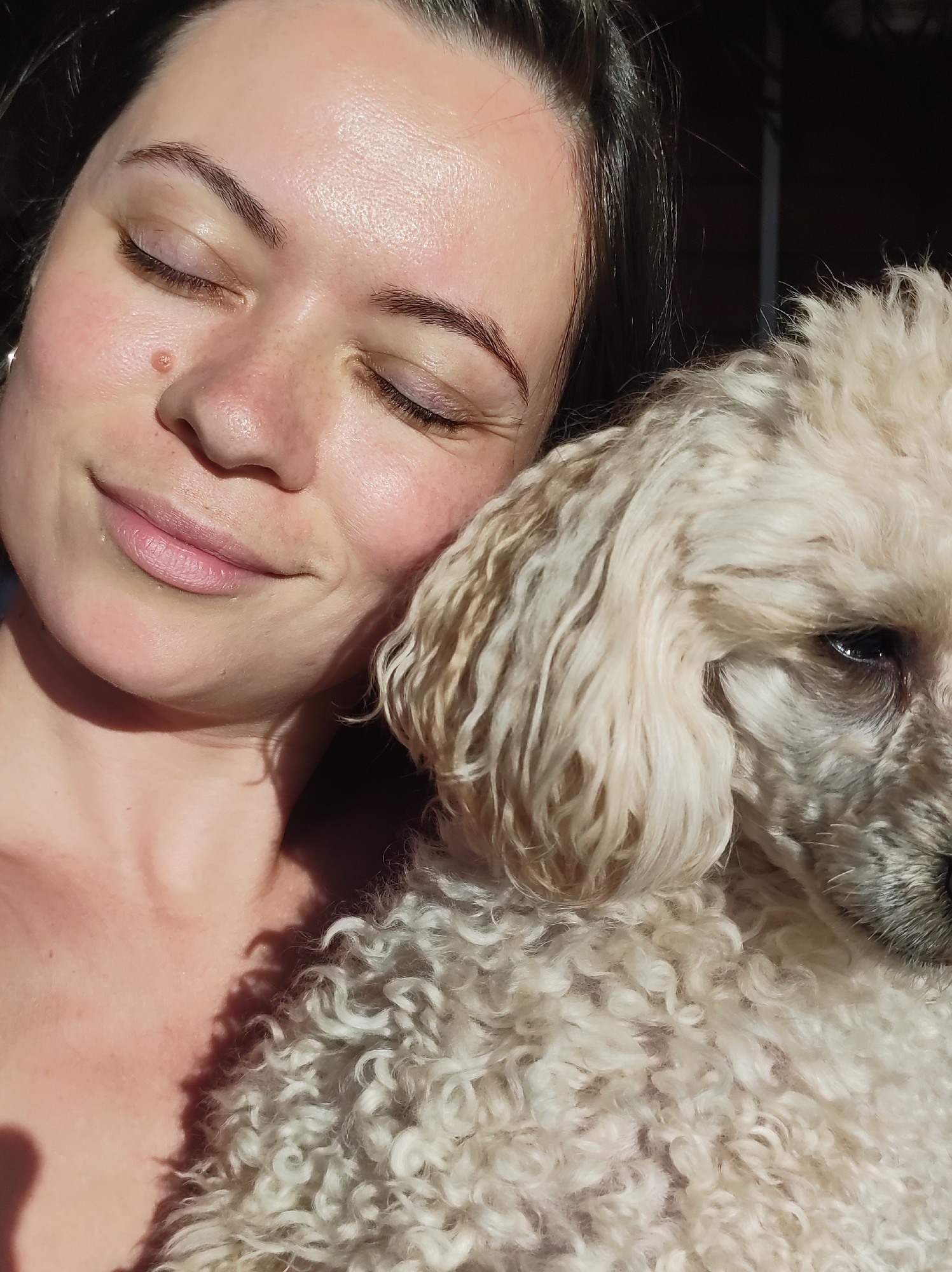 A woman with dark hair, her eyes closed and a slight smile on her lips, is cuddling a white poodle. The woman has a mole on her left cheek and is outdoors in natural light. The dog is primarily white with some gray around its muzzle and one eye visible. The location is in Фоки, Russia.