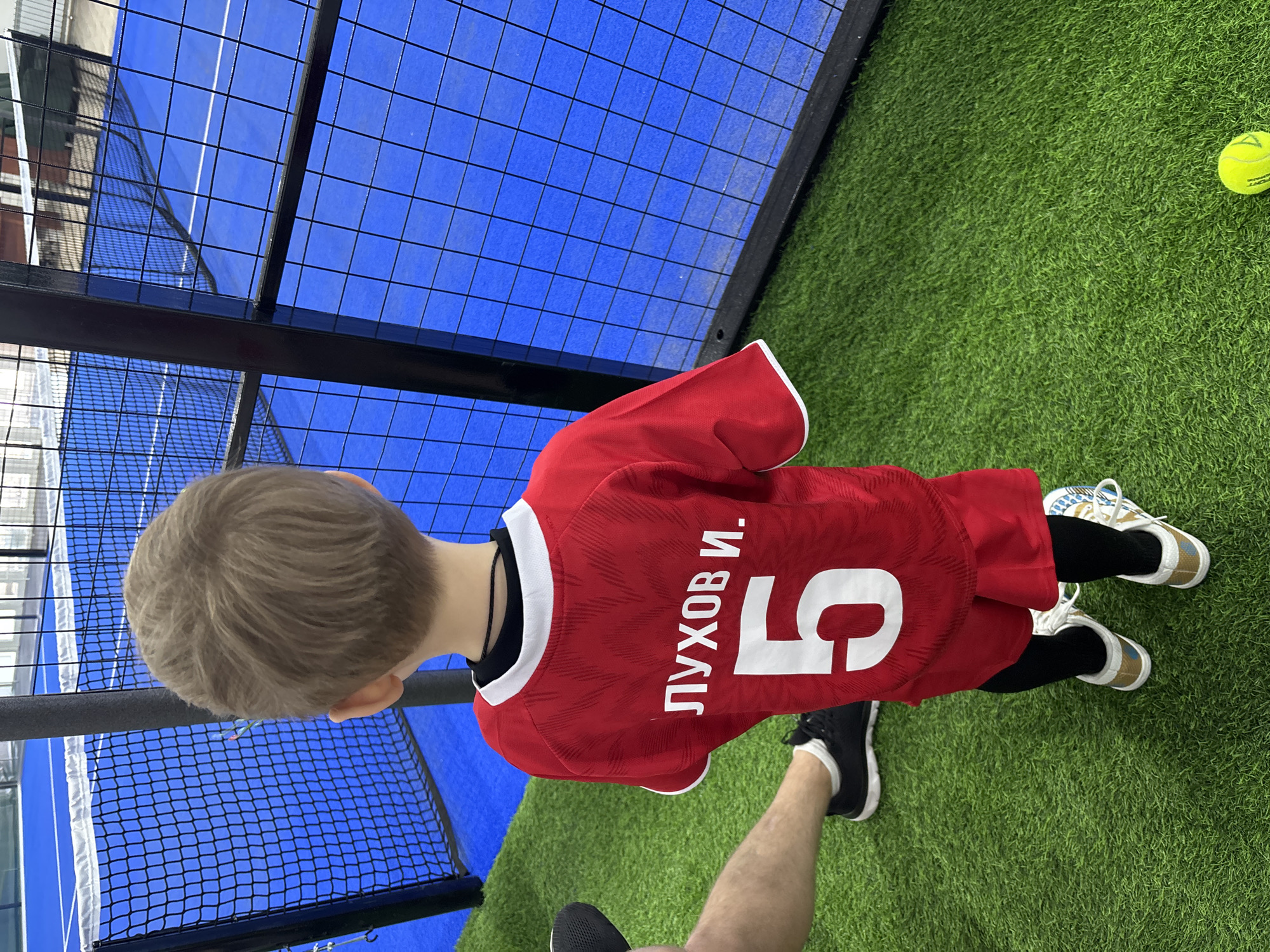 A young boy wearing a red soccer jersey with the name "Лухов И." and the number "5" is standing on artificial grass next to a blue tennis court with black mesh fencing. A tennis ball is visible on the grass. An adult's leg and foot wearing a black shoe are also visible. The location is likely in the city of Chaikovsky, Russia, based on the Cyrillic lettering on the jersey.