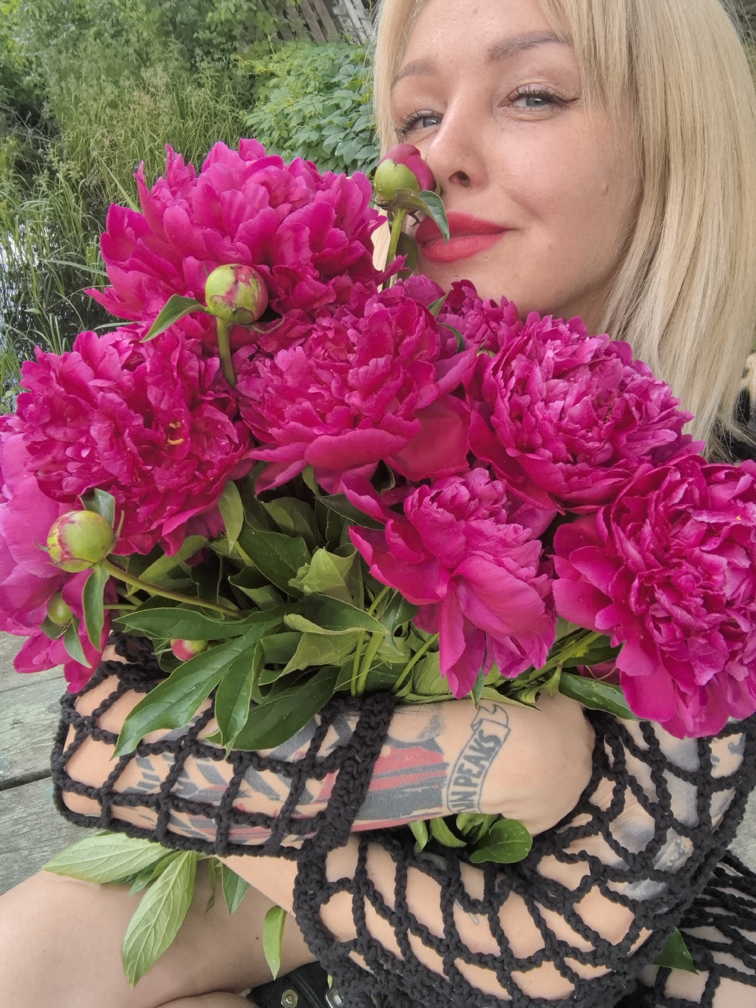 A Caucasian woman with blonde hair and red lipstick is holding a large bouquet of vibrant pink peonies. She is wearing a black crocheted garment that covers her arms and chest. Tattoos are visible on her forearms, one of which reads "Twin Peaks." The woman is outdoors, with lush green foliage and trees in the background, suggesting a natural or park-like setting. The ground beneath her appears to be made of wooden planks, possibly a deck or pier. The overall impression is a candid portrait of a woman enjoying flowers in an outdoor environment. The image does not provide any specific information that would allow it to be definitively located in Noviy, Russia.