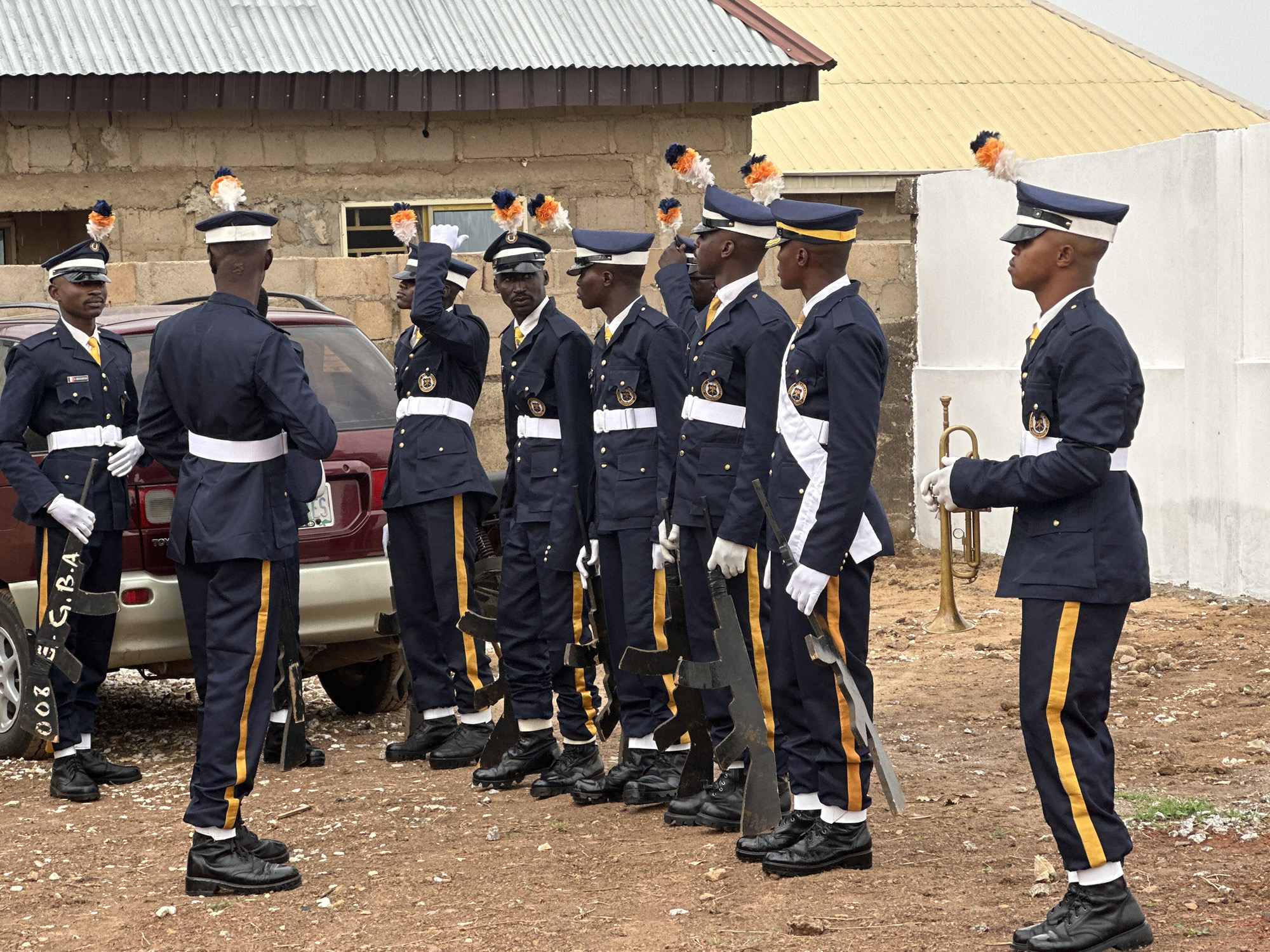 A line of eight individuals in dark blue uniforms with gold trim and matching hats with orange and white plumes stand at attention in Goni Gara, Nigeria. Seven of them hold what appear to be decorative rifles with white-gloved hands, and one holds a trumpet. The uniforms include white belts and white gloves. The rifles have the inscription "002 G. GBA". They stand on a brown, grassy area in front of a light brick building and a white wall. A maroon SUV is visible behind the first few individuals.