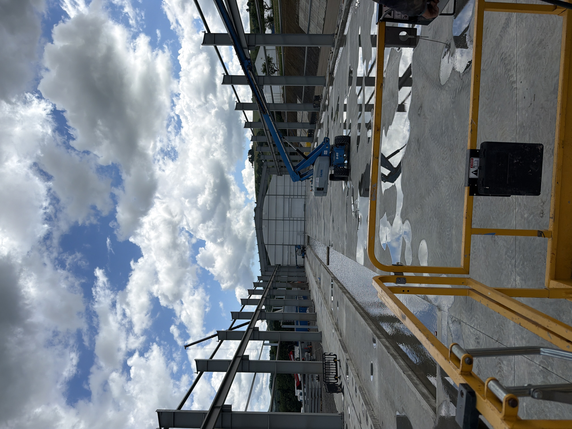This is a full, eye-level, outdoor shot of a construction site in Stróża, Poland. The image captures a large, partially constructed steel building with a light-colored, curved roof. A blue boom lift is positioned in the middle ground, with its arm extended upwards, resting on one of the building's steel beams. The ground is concrete, with puddles of water reflecting the sky and clouds. In the foreground, a yellow metal safety railing is visible, partially obscuring the view. The sky is filled with dramatic, white and gray cumulus clouds against a bright blue backdrop. In the distance, green hills and trees are visible. The scene appears to be a typical construction project underway on a cloudy day.