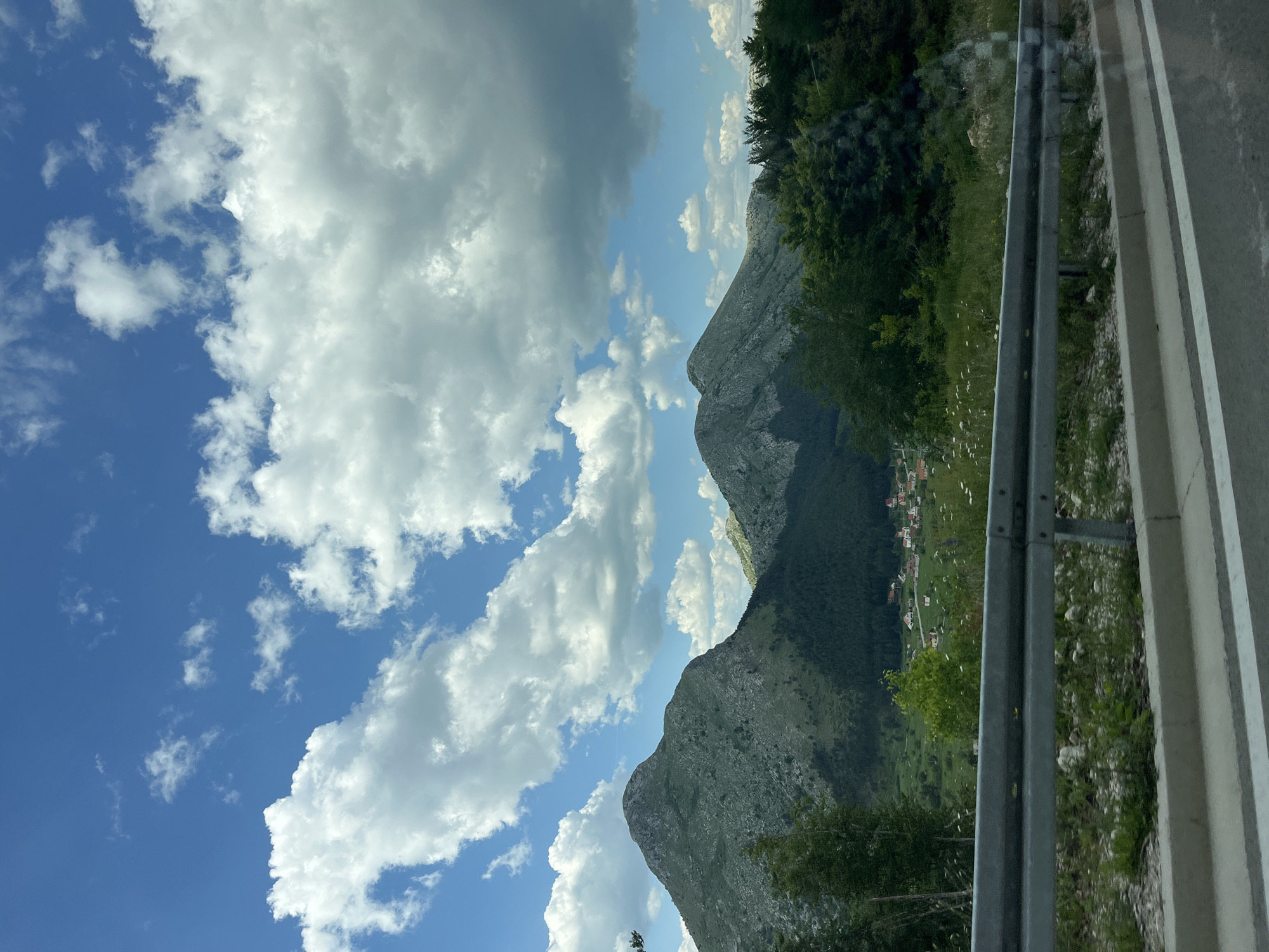 The image, taken at an angle, captures a mountainscape under a blue sky with scattered white clouds. The mountains are rocky and partly covered with green vegetation. A small village or town, presumably Mali Bostur, is visible nestled amidst the greenery at the base of the mountains. A metal guardrail runs along the edge of a road in the foreground. The scene is generally bright and appears to have been captured during daylight hours.