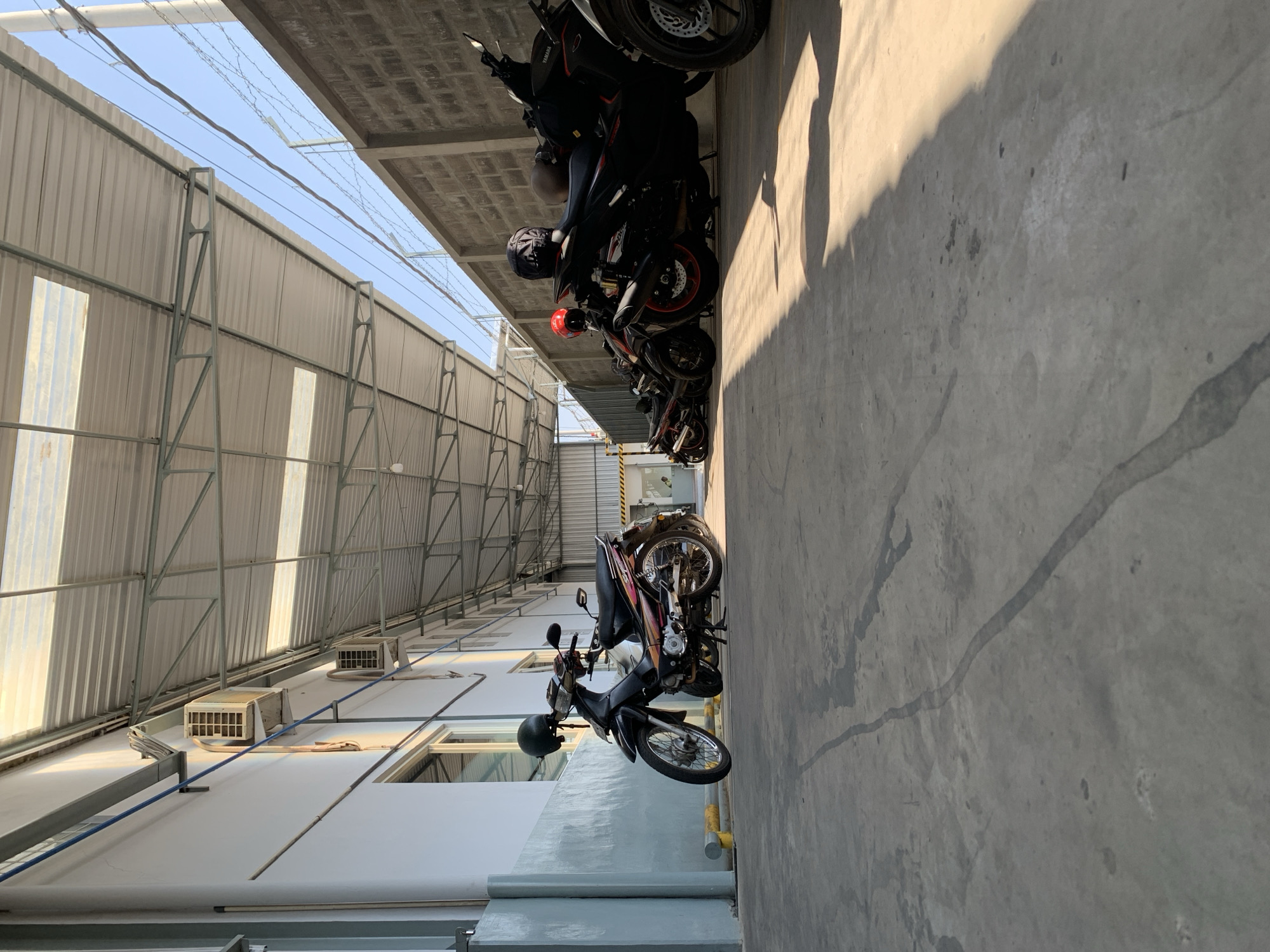 The image depicts a covered outdoor parking area, likely in Karangploso, Indonesia, adjacent to a building with a white exterior and visible air conditioning units mounted on the wall. The roof of the parking area is constructed with corrugated metal and translucent panels, supported by a metal frame. A concrete floor with some water stains and a prominent line of discoloration runs through the foreground. Several motorcycles are parked in the area, with one black and silver scooter in the foreground, and a row of darker motorcycles, some with helmets on them, extending further into the background. A brick wall with barbed wire on top forms the boundary on the right side of the parking area. The scene is illuminated by natural sunlight, casting shadows on the concrete floor. There are no people visible in the image.