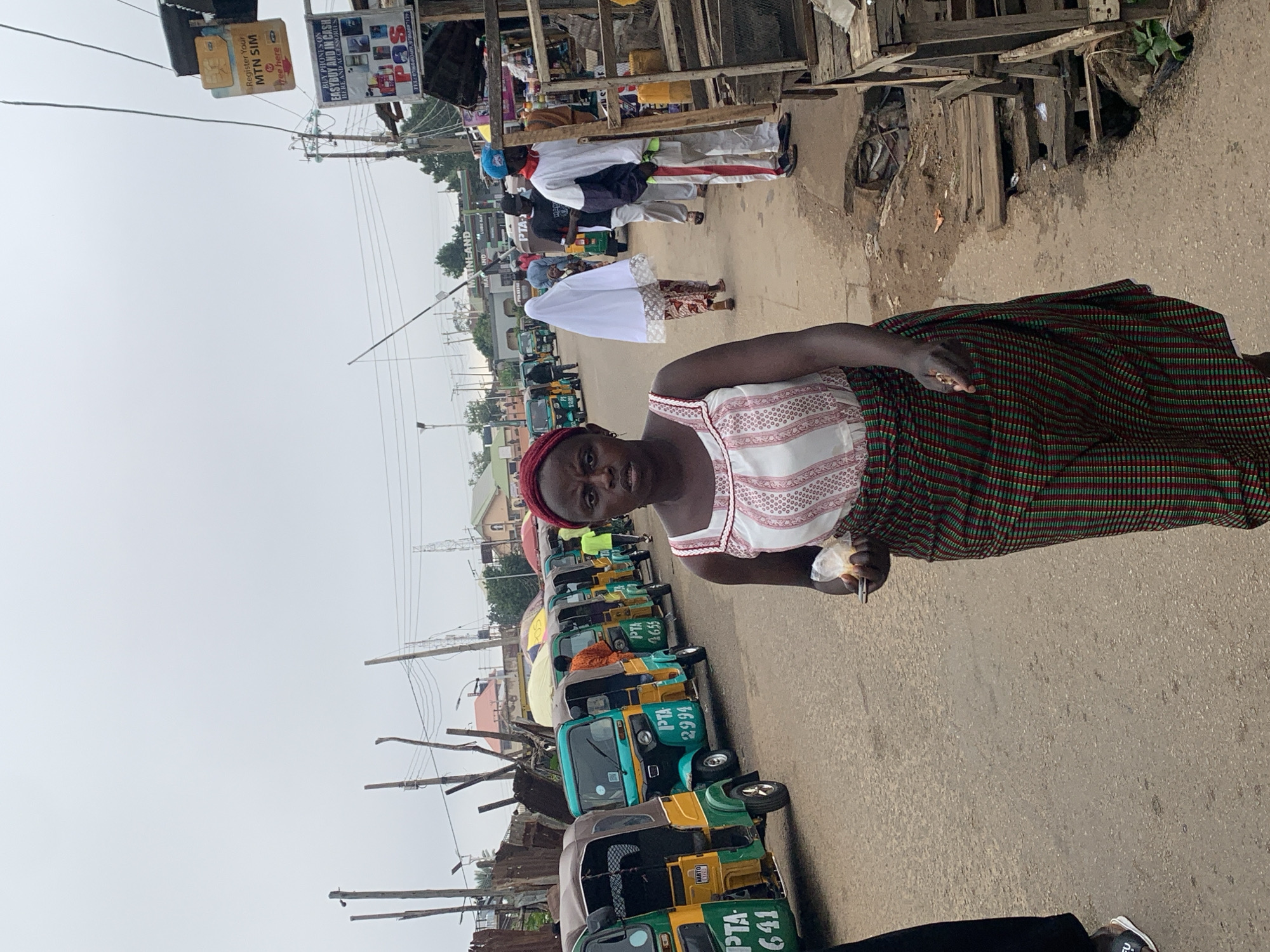 This media file depicts a street scene in Jos, Nigeria, likely during the daytime. The foreground features a woman wearing a red headscarf, a white and red patterned top, and a green and red striped wrap skirt. She is holding a small white object, possibly food, and a phone in her hands.
Behind her, a row of colorful auto-rickshaws, commonly known as "Kekes," are parked along the roadside. Some of these vehicles have visible text, including "PTA 641," "IPTA 2994," and "PTA 4655." The scene suggests a bustling urban environment, possibly a marketplace or a transportation hub.
Further in the background, other individuals can be seen, some in traditional attire and others in more casual clothing. A signpost advertising "MTN SIM" and "BUY PHONES ON EASYBUY AND IN CASH" is also visible, indicating commercial activity. The overall atmosphere appears to be that of a lively street scene, with people and vehicles creating a sense of daily life and commerce. The weather appears overcast, contributing to a diffused light.