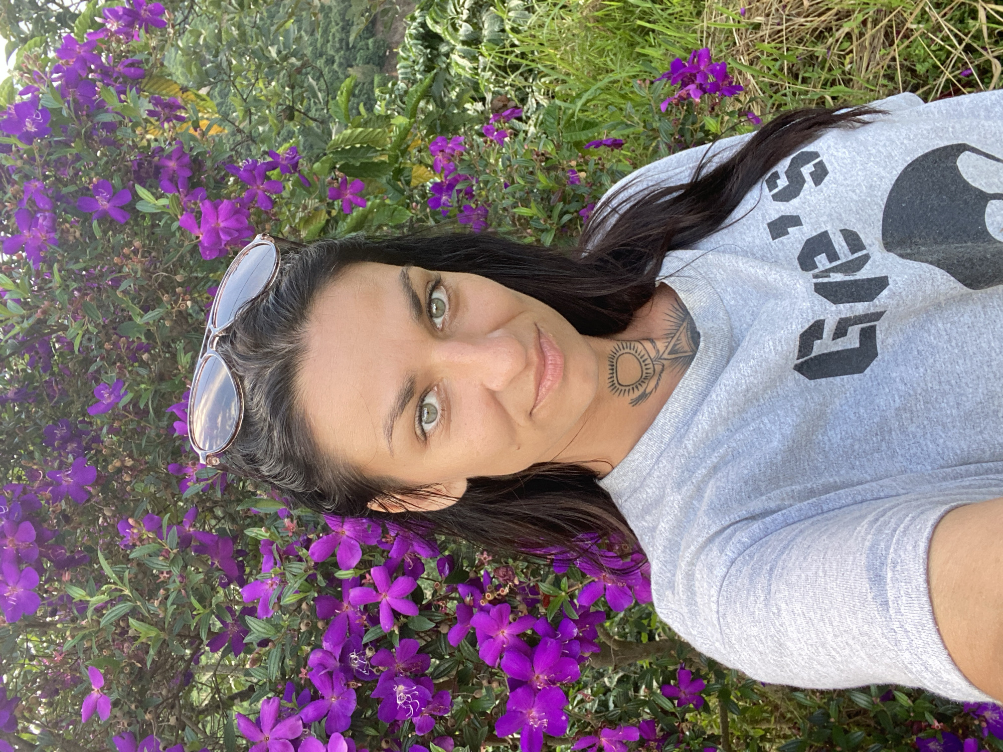 A young adult Caucasian woman with dark hair and green eyes is pictured in a selfie. She is wearing a light grey t-shirt with a graphic design and has a tattoo on her neck. Sunglasses are perched on her head. She is standing outdoors in front of a bush with vibrant purple flowers. The background consists of green foliage and grass, suggesting a natural environment. The specific location in Thôn 3, Vietnam is not discernible from the image.