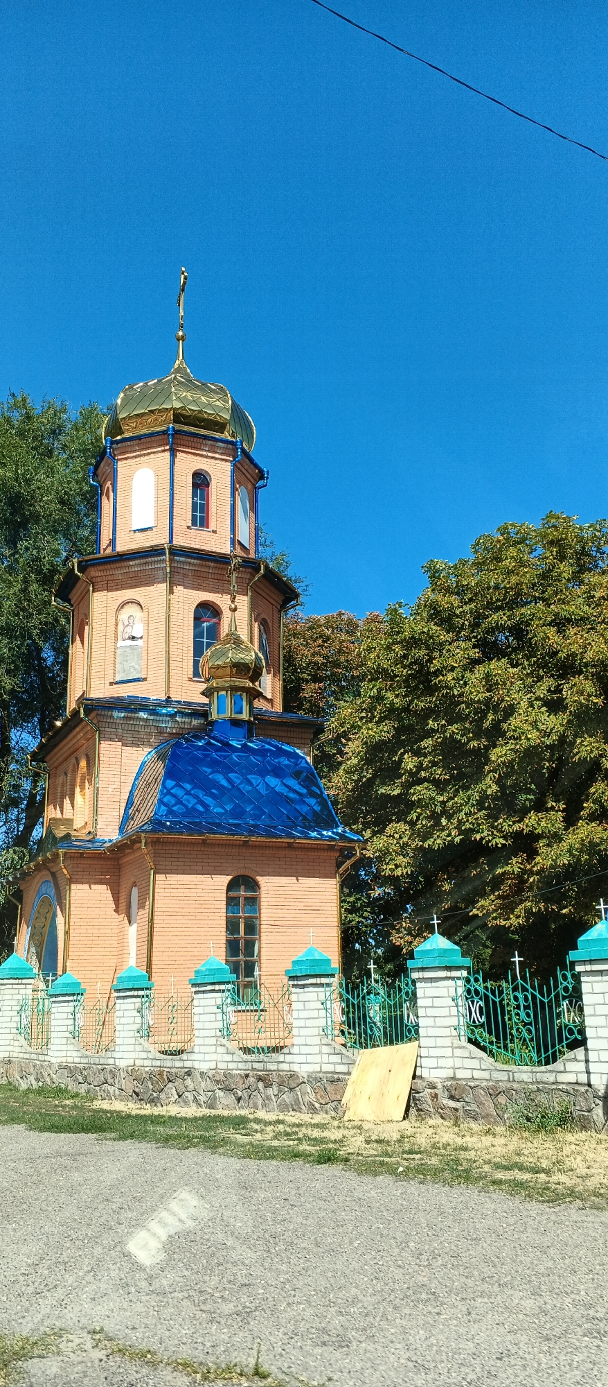 The image shows a small, brick church with a golden dome and a blue tiled roof, located in the city of Smila, Ukraine. The church has a bell tower with multiple levels and windows. A fence made of white brick with turquoise accents and green metalwork surrounds the church property. Trees with green and brown foliage are visible behind the church and fence. The sky is a clear, bright blue. There is a wire visible in the upper portion of the image, and a piece of plywood leaning against the fence in the foreground. The ground in front of the fence is covered with gravel and some dry grass. There are no people or animals visible in the image.