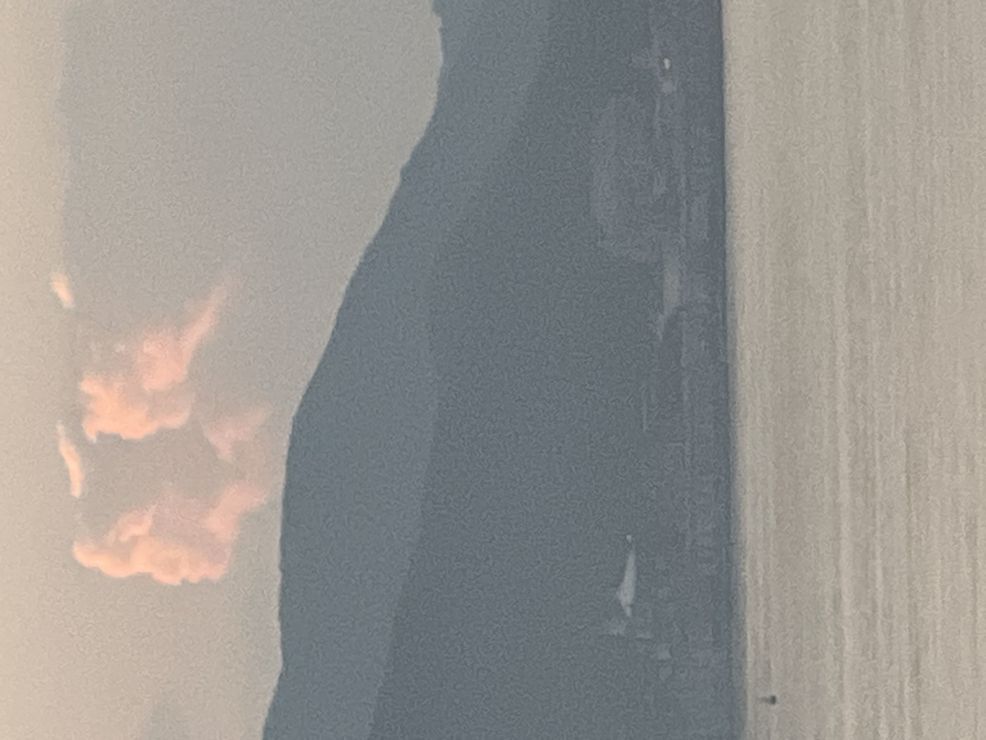 The image depicts a hazy sky with a silhouette of a mountain range in the background. Above the mountains, clouds are visible, with one cloud formation appearing to resemble a human profile with rosy hues. The foreground shows a body of water, likely the sea, with a faint shimmer on its surface. In the distance, along the base of the mountain range, a city skyline is discernible, with buildings and structures. A small boat is visible in the water in the lower left portion of the image. The overall scene suggests an outdoor setting, possibly during dawn or dusk, given the soft, diffused light. The location is identified as Nha Trang, Vietnam, implying the mountain range and sea are characteristic of the coastal city's geography.