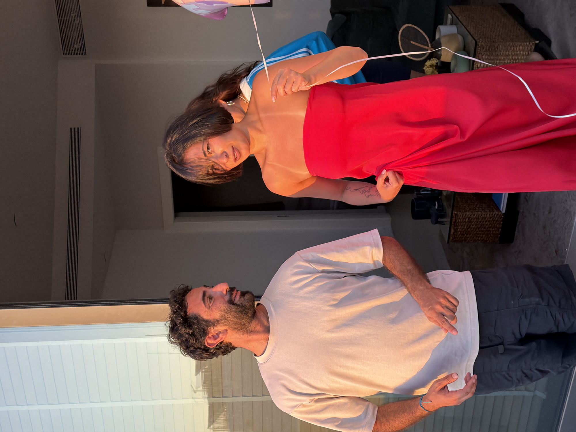 A man with dark curly hair and a beard is standing to the left of the frame, wearing a light-colored t-shirt and dark shorts. He is looking to his right. To his right, a woman with dark hair is wearing a strapless red dress. She is holding a white ribbon and looking at the camera. Behind her, another person in a blue jacket with white stripes is partially visible. The scene appears to be on a balcony or patio, with a glass door or window to the left reflecting sunlight. Indoor furniture, including a coffee table with decorative items, is visible in the background. The lighting suggests it is either sunrise or sunset, with a warm glow on the subjects. The location is likely a residential building in Tel Aviv, Israel, given the architecture and outdoor setting often found in the city.