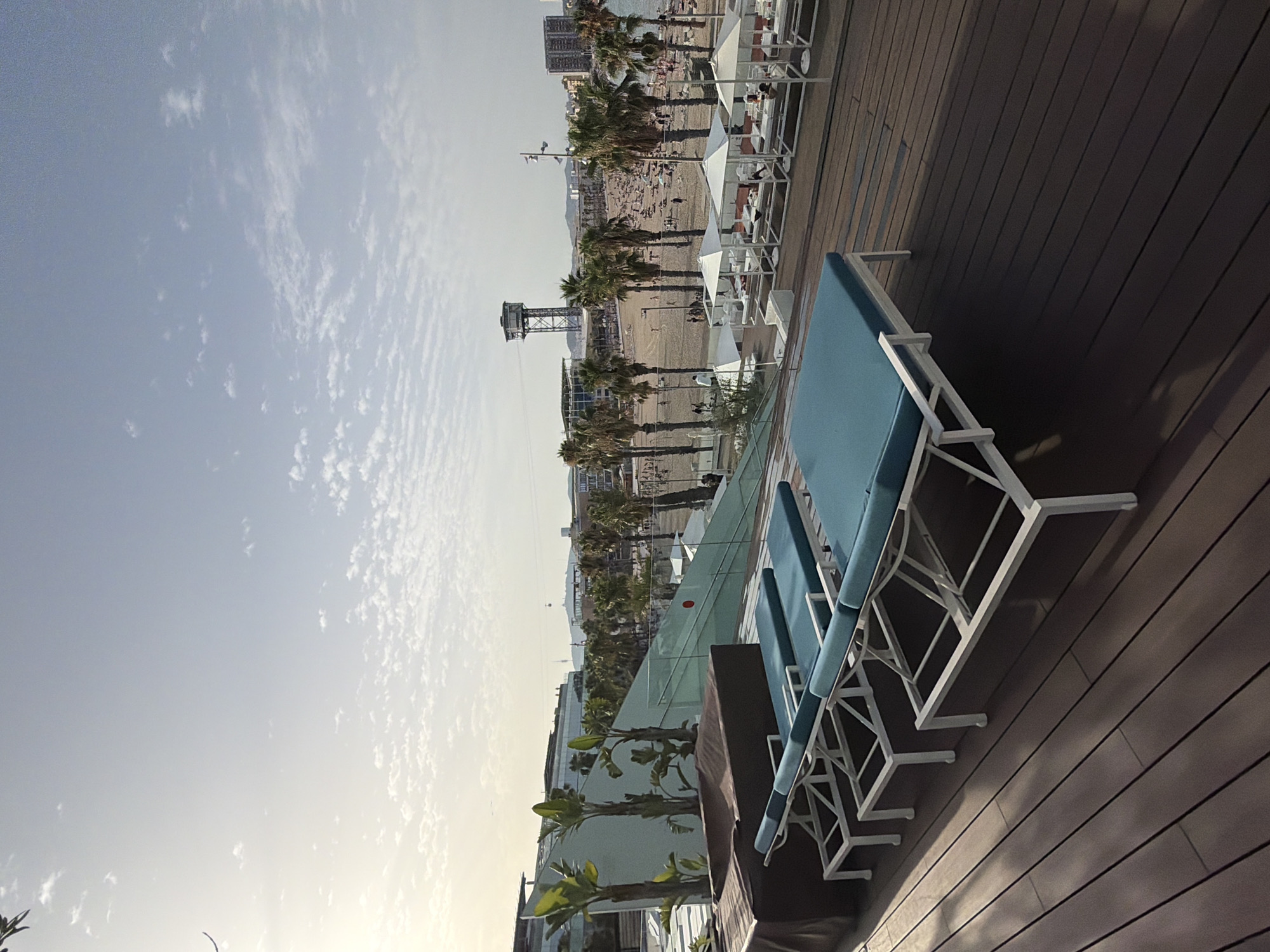 The image depicts a rooftop terrace overlooking Barceloneta Beach in Barcelona, Spain. In the foreground, several light-colored lounge chairs with blue cushions are arranged on a dark wooden deck. Behind the lounge chairs, a modern building with a slanted glass facade and tropical plants is visible. Beyond this, a crowded sandy beach stretches along the coastline, dotted with palm trees. In the background, various buildings and a distinctive industrial tower are visible against a partly cloudy sky. The scene suggests a leisure setting with a view of the Mediterranean Sea and the city.