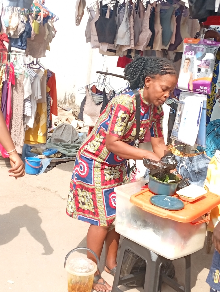 A woman in a patterned dress stands at a makeshift food stall. She's packing food into containers atop a plastic box on a stool. Behind her, clothing items hang on racks, indicating an outdoor market or street vending environment. A bucket with a beverage sits on the ground, and another container is visible on the food stall. Other people are faintly visible.