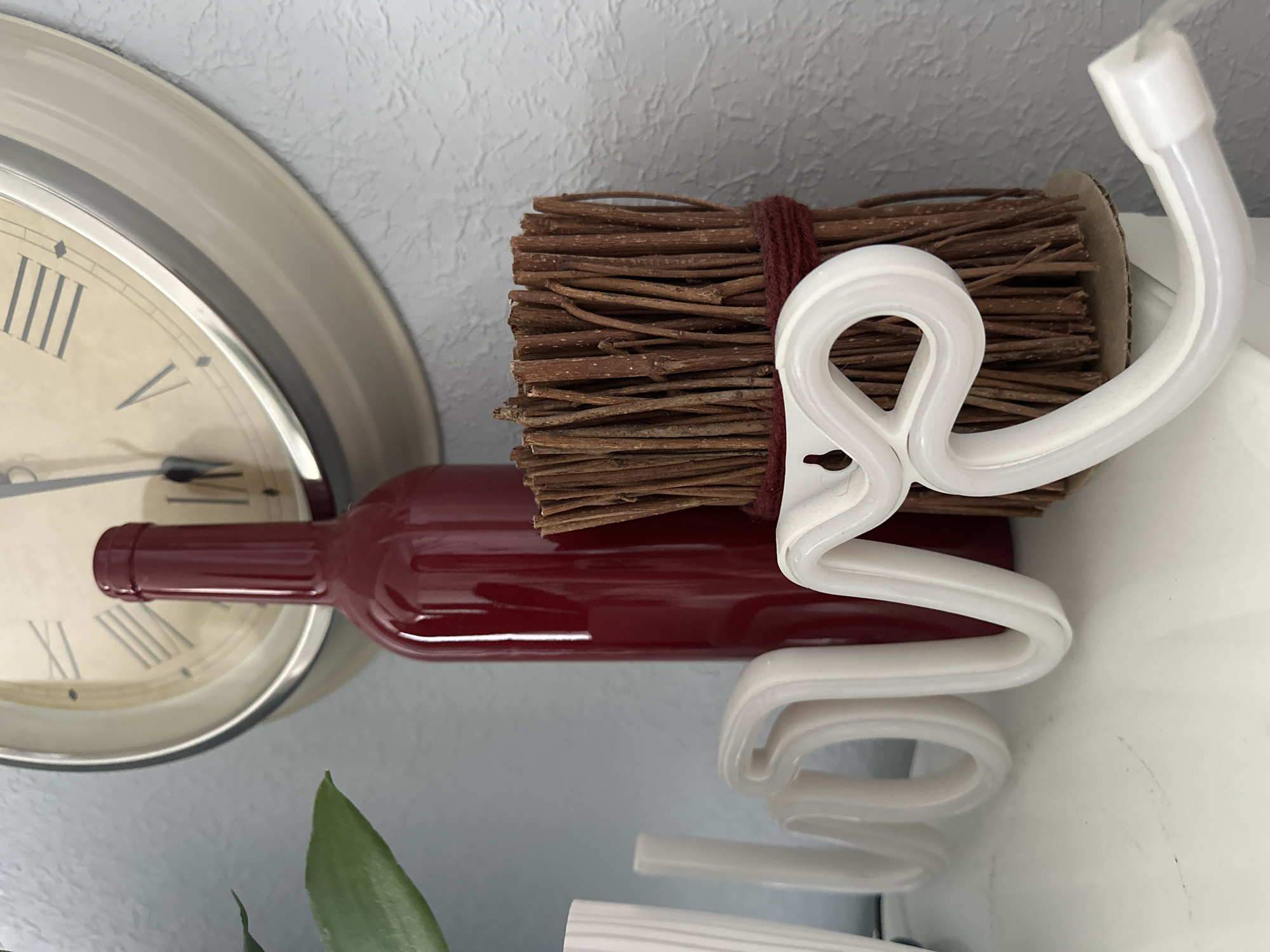 A close-up, eye-level, medium shot captures a decorative arrangement on a white shelf against a textured light blue wall. Dominating the center is a tall, deep red glass bottle. To its right, a bundle of thin, brown twigs is tied with dark red twine, resting on a cardboard base. In front of these, a white neon sign spelling "love" curves across the foreground, with the 'l' and 'o' on the left and the 'v' and 'e' extending to the right. Behind the bottle, a portion of a large, cream-colored wall clock with Roman numerals is visible. A green leaf from an unseen plant is positioned to the left of the clock. The lighting appears to be natural, casting soft shadows. The image does not depict any people or actions. The location is not identifiable from the provided image.