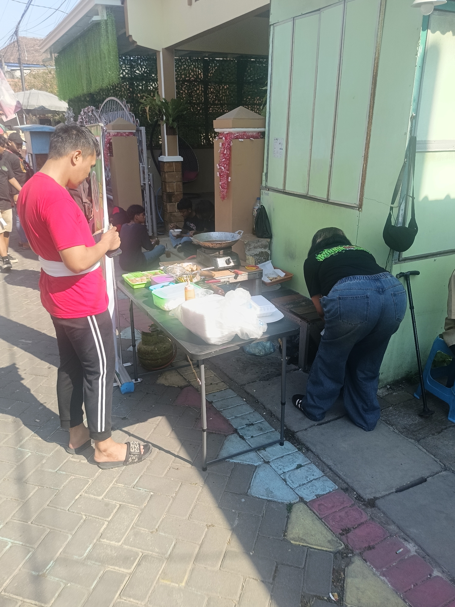 In the city of Gedangan, Indonesia, on a sunny day, a man in a red t-shirt and black pants with white stripes stands facing left, holding a smartphone. He is positioned on a brick-paved sidewalk. To his right, a black folding table is set up. On the table are various food items in containers, white styrofoam boxes, and a gas cylinder. A woman in a black t-shirt with a green logo and loose blue jeans is bending over the table, engaged with the items. Behind her, a walking stick leans against a light green wall. Further back, another table is visible with cooking utensils, including a wok on a stove. In the background, the scene shows a building with a light-colored facade and green accents, with some decorative greenery hanging from above. Adjacent to the building is a gate and other individuals are partially visible in the background. The overall environment suggests an outdoor food stall or market setting.