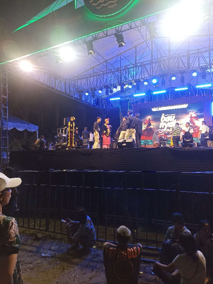 This image depicts an outdoor night event centered around a large, brightly lit stage in Kecamatan Pujut, Indonesia. The stage is a robust structure built with metal trussing, supporting numerous blue and white stage lights, as well as green accent lighting along its upper frame. At the very top, a banner or screen displays a circular logo with wavy lines.
On the stage, several people are visible. Two individuals stand out due to their traditional attire: one on the left wears an elaborate dark costume with gold embellishments and a tall headdress, while another near the center is dressed in reddish-pink traditional garments and headwear. Other individuals in casual clothing are also present on the stage, some seemingly involved in preparations or as part of the event crew. Behind the performers, a large LED screen displays dynamic, colorful graphics with visible text, including parts of words like "bau..." and "NYANYI," indicating a performance or cultural show.
In the foreground, a metal barrier separates the stage from the audience area. Below this barrier, several spectators are sitting directly on the dirt ground. One person is engrossed with their mobile phone, while another, with short blonde hair, wears a black t-shirt featuring a prominent circular logo on the back. A person wearing a white cap is partially visible on the far left. The scene is illuminated almost entirely by the stage lights, creating a bright focal point against the dark night sky, suggesting an evening or nighttime cultural performance or concert.