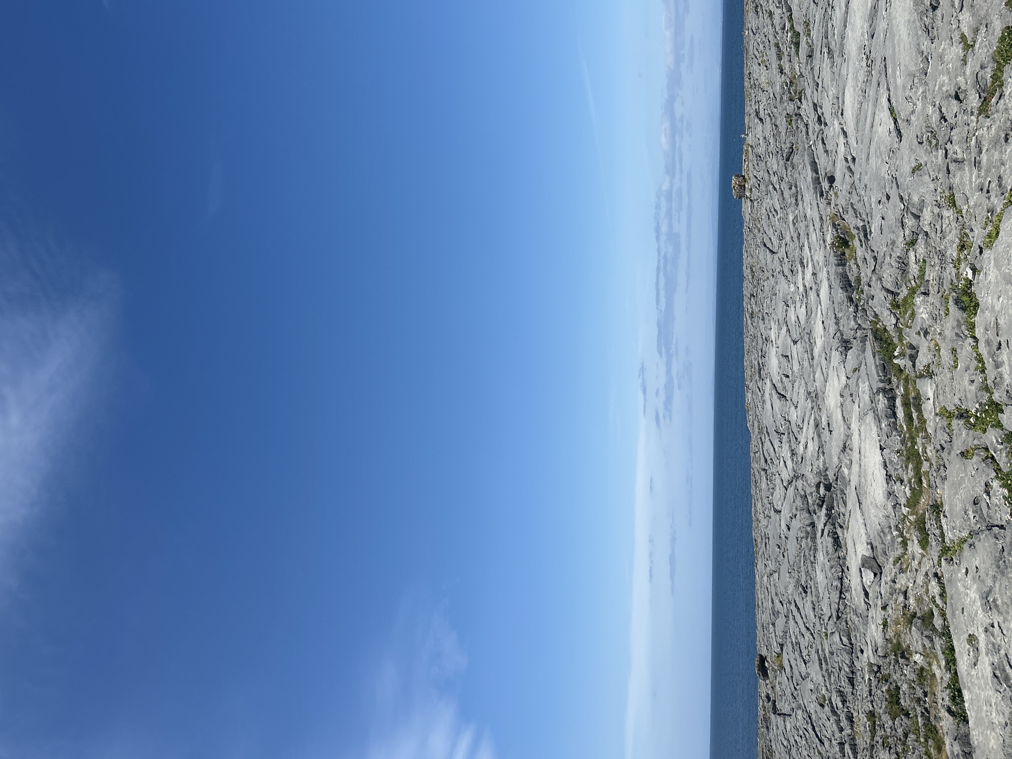 A wide shot shows a rocky coastline under a bright blue sky in Ireland. The foreground is dominated by a vast expanse of grey, weathered limestone, characteristic of the Burren region, with sparse green vegetation pushing through cracks. In the mid-ground, the deep blue sea stretches to the horizon, meeting a hazy distant shoreline. A few wisps of white clouds are scattered across the upper portion of the sky. A solitary, rough-hewn stone structure stands on the rocky terrain near the water's edge. The scene evokes a sense of wild, natural beauty.