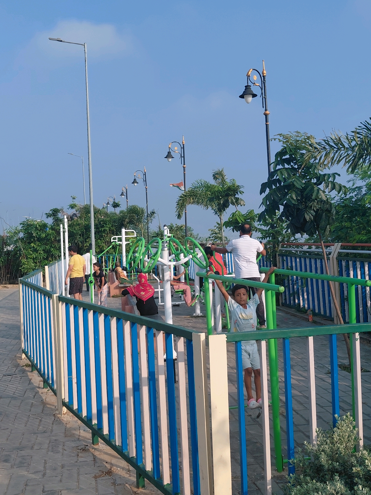 This is a photograph taken in the city of Patna, India, likely at an outdoor fitness park. The image features a bright, sunny day with a clear blue sky. In the foreground, there is a colorful fence with blue, white, and green vertical bars. Beyond the fence, there are several pieces of outdoor exercise equipment, predominantly green and white. Several individuals are visible in the park, some using the equipment and others observing. A man in a yellow t-shirt and shorts stands near the fence, and a young girl in a black t-shirt and patterned leggings is nearby. Another child, a boy in a light blue t-shirt and grey shorts, is actively engaged with the exercise equipment, holding onto a horizontal bar. Further back, a man in a striped white t-shirt and dark shorts is also using the equipment. A woman in a red headscarf and black top is seated on a bench. The park is surrounded by lush green trees and vegetation, and several streetlights are visible, including a tall, modern one in the background and more ornate, decorative ones closer to the park. The ground is paved with interlocking stones.