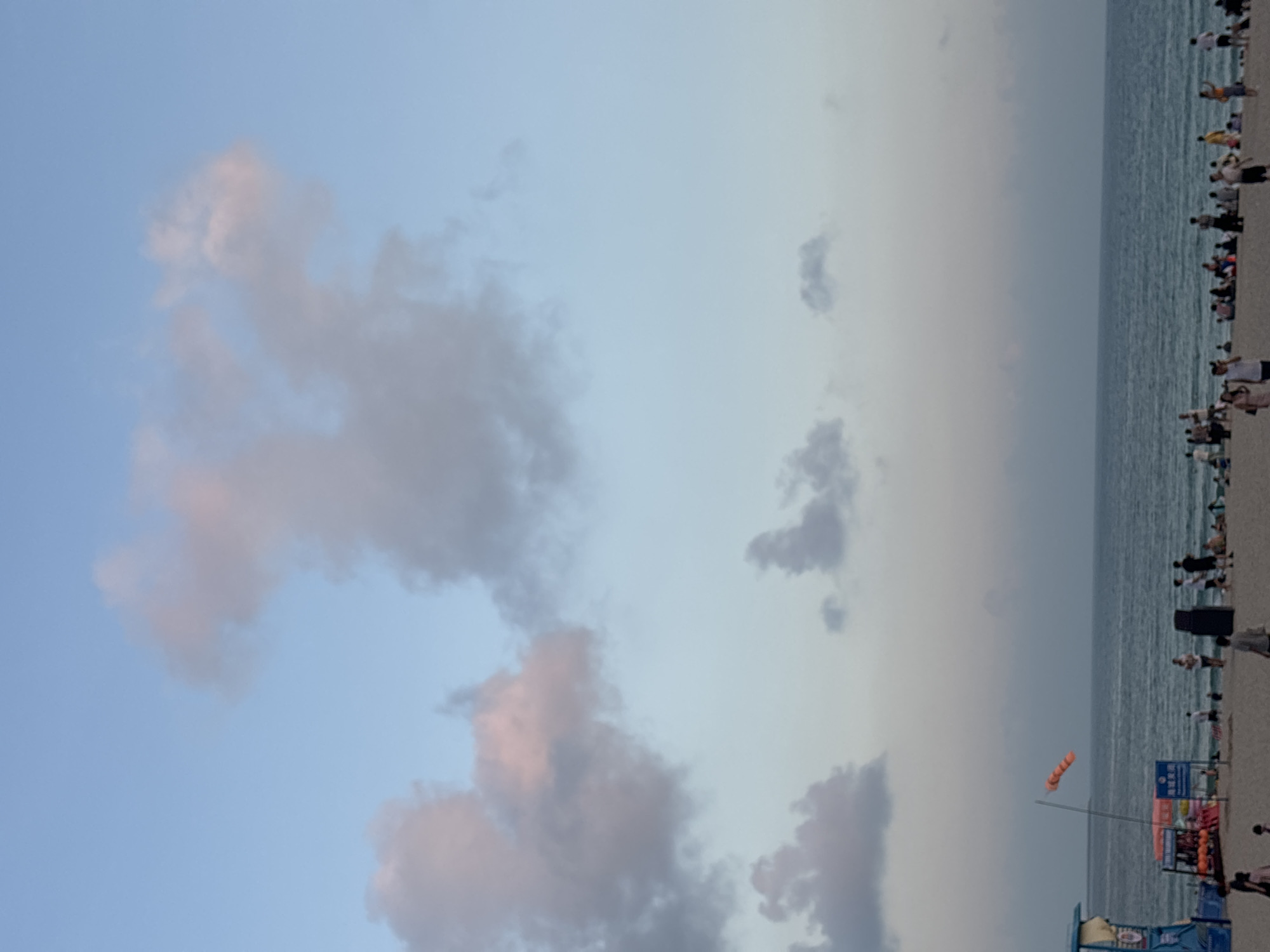 The image depicts a beach scene with a calm ocean under a sky with scattered clouds. The sky transitions from a light blue at the top to a soft gradient of pink and grey near the horizon. Several people are visible on the sandy beach, some walking, others sitting, and a few appear to be interacting. In the foreground, on the left side, a blue lifeguard station is partially visible, with some people gathered around it. A windsock, colored orange and white, is mounted on a pole near the water's edge, indicating light wind. The overall atmosphere suggests a peaceful evening on a populated beach. This scene is located in Jiyang District, China.