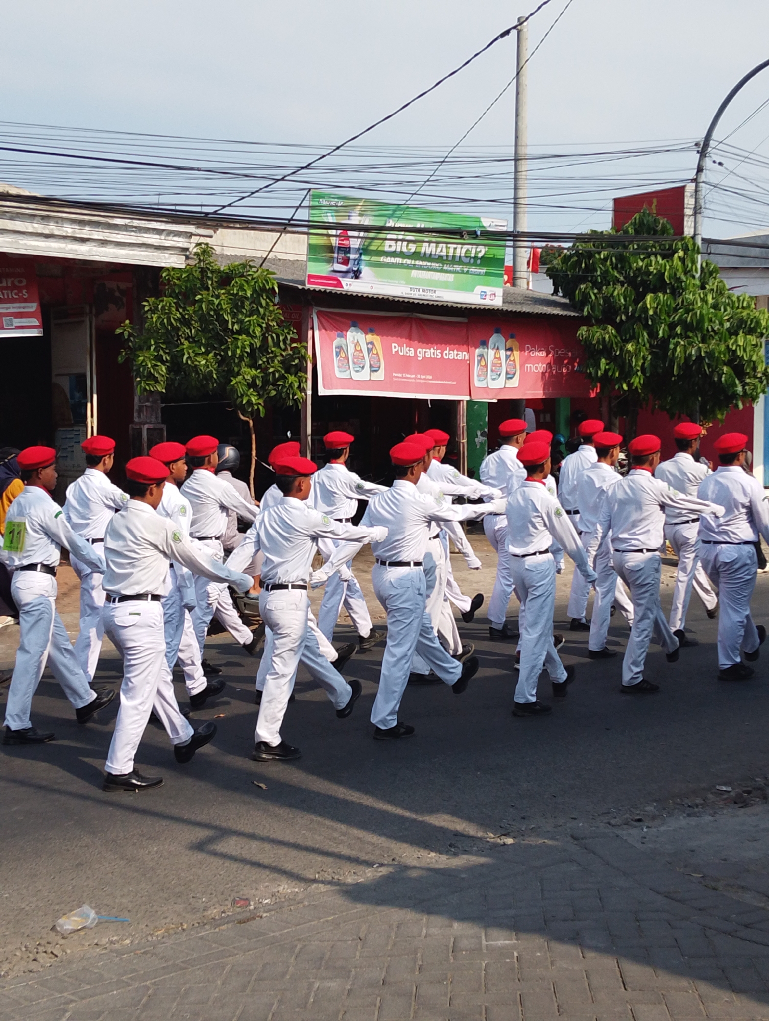 In Manaruwi, Indonesia, a group of uniformed individuals in white shirts, white pants, and red berets are marching in formation on a paved road. They appear to be participating in a parade or a similar organized event. The background features buildings with red awnings and green trees, suggesting a commercial or residential area within the city. Overhead, a network of power lines crisscrosses the sky. The lighting indicates it is daytime, likely morning or afternoon.