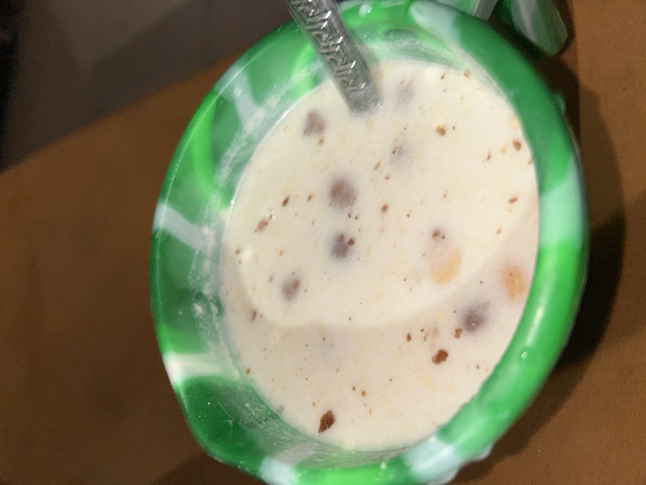 A close-up, top-down view depicts a bowl containing a liquid food substance within an indoor setting in Egbe, Nigeria. The bowl is circular, made of a green and white marbled material, possibly silicone. It holds a light, creamy-colored liquid in which various solid components are suspended: numerous dark brown, irregularly shaped spherical particles, smaller lighter brown/orange fragments, and fine dark specks. A silver-colored spoon, featuring a decorative pattern on its handle, is partially submerged in the liquid on the right side of the bowl. The bowl rests on an out-of-focus, dark brown surface, likely a table. No individuals or actions are visible.