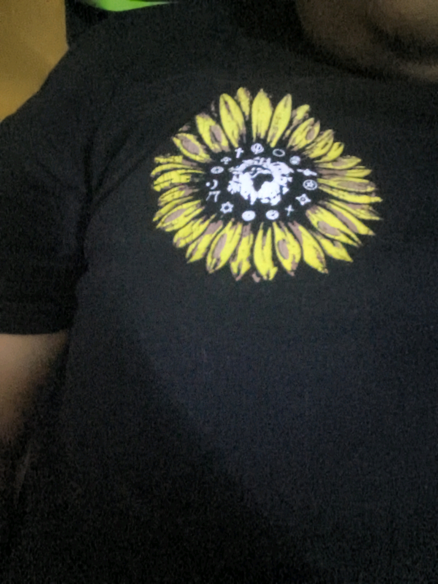 A close-up, full-body photograph shows a person wearing a black t-shirt with a sunflower graphic design on the chest. The sunflower is depicted in yellow and brown, with various symbols embedded within its petals and center. The background is dark and out of focus, offering no discernible details about the environment or location. The person's face is not visible, and only a portion of their upper body is in the frame. The lighting is dim, with some ambient light illuminating the t-shirt.