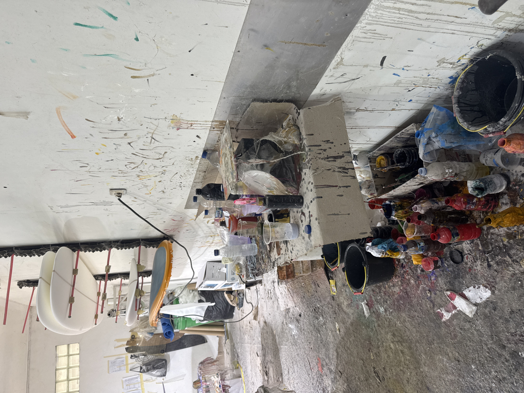 The image shows an artist's studio, likely in Denpasar, Indonesia, though no specific landmarks are visible. The room is cluttered with painting supplies. Surfaces are covered in paint splatters of various colors. Numerous paint bottles, buckets, and plastic containers are scattered across the floor and on shelves. Some white rectangular objects, possibly canvases, hang from the ceiling. The environment suggests a workspace dedicated to painting. No people are visible.